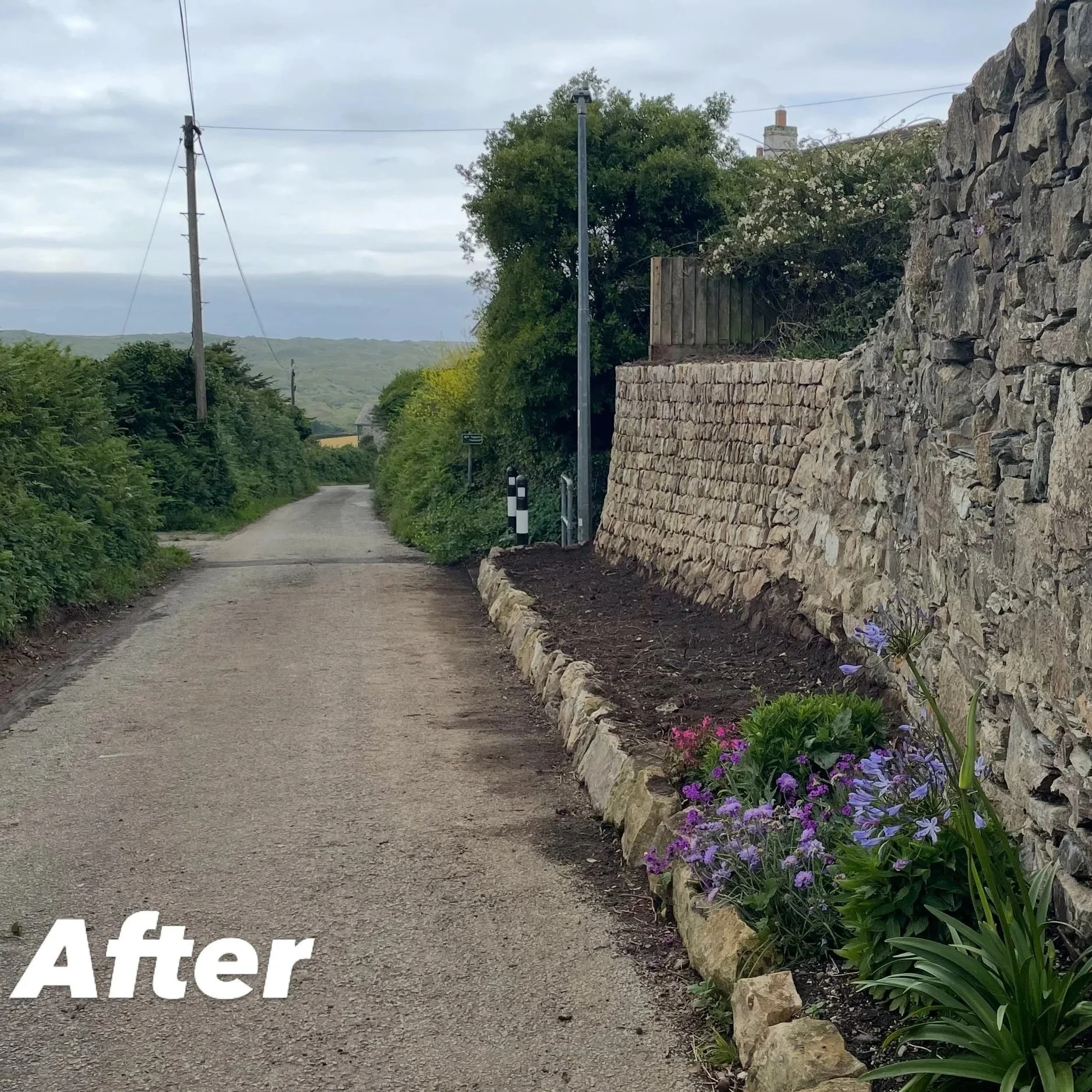 A gravel pathway lined with a stone wall on the right side and flowering plants, with a background of trees and rolling hills, under a cloudy sky.