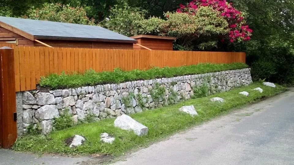 A wooden fence and a stone retaining wall with green plants on top, alongside a gravel road with large rocks at its base and in the grass.