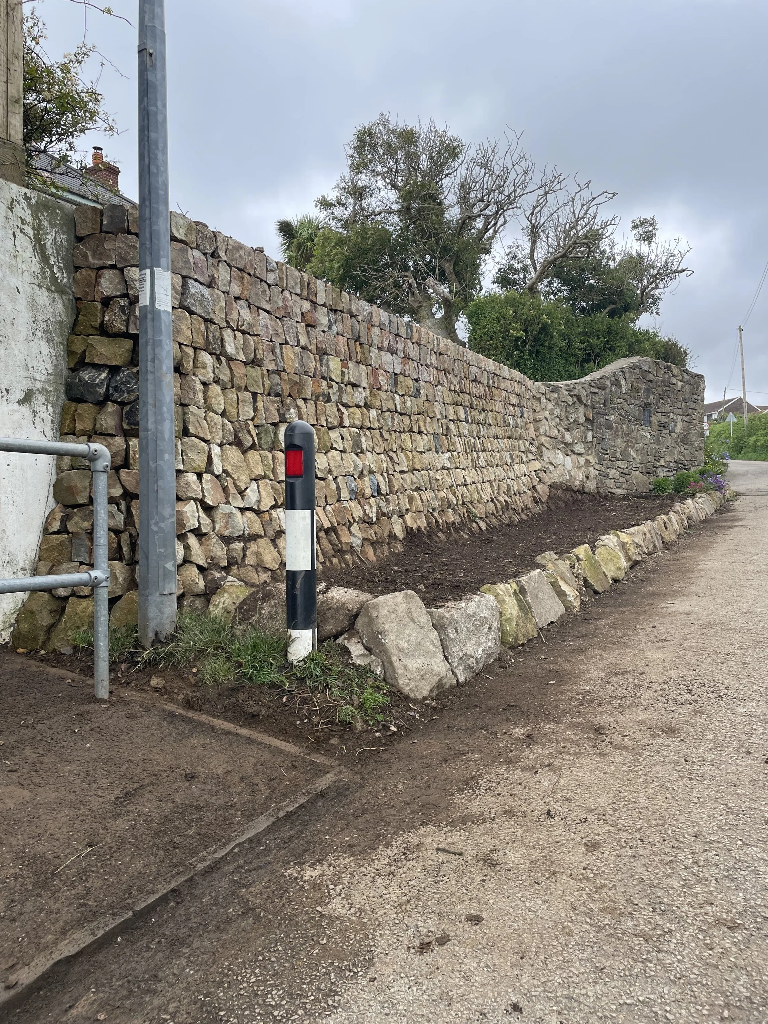 A stone retaining wall with a curved section, beside a dirt pathway, and a large leafless tree with some greenery on top in the background. There are traffic posts and a metal railing near the pathway, under a cloudy sky.