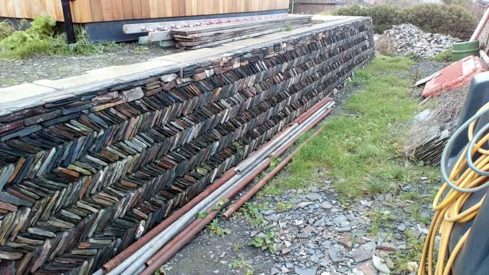 A long stack of firewood split and arranged neatly outside a building, with some pipes and construction materials nearby on a grassy and gravelly ground.