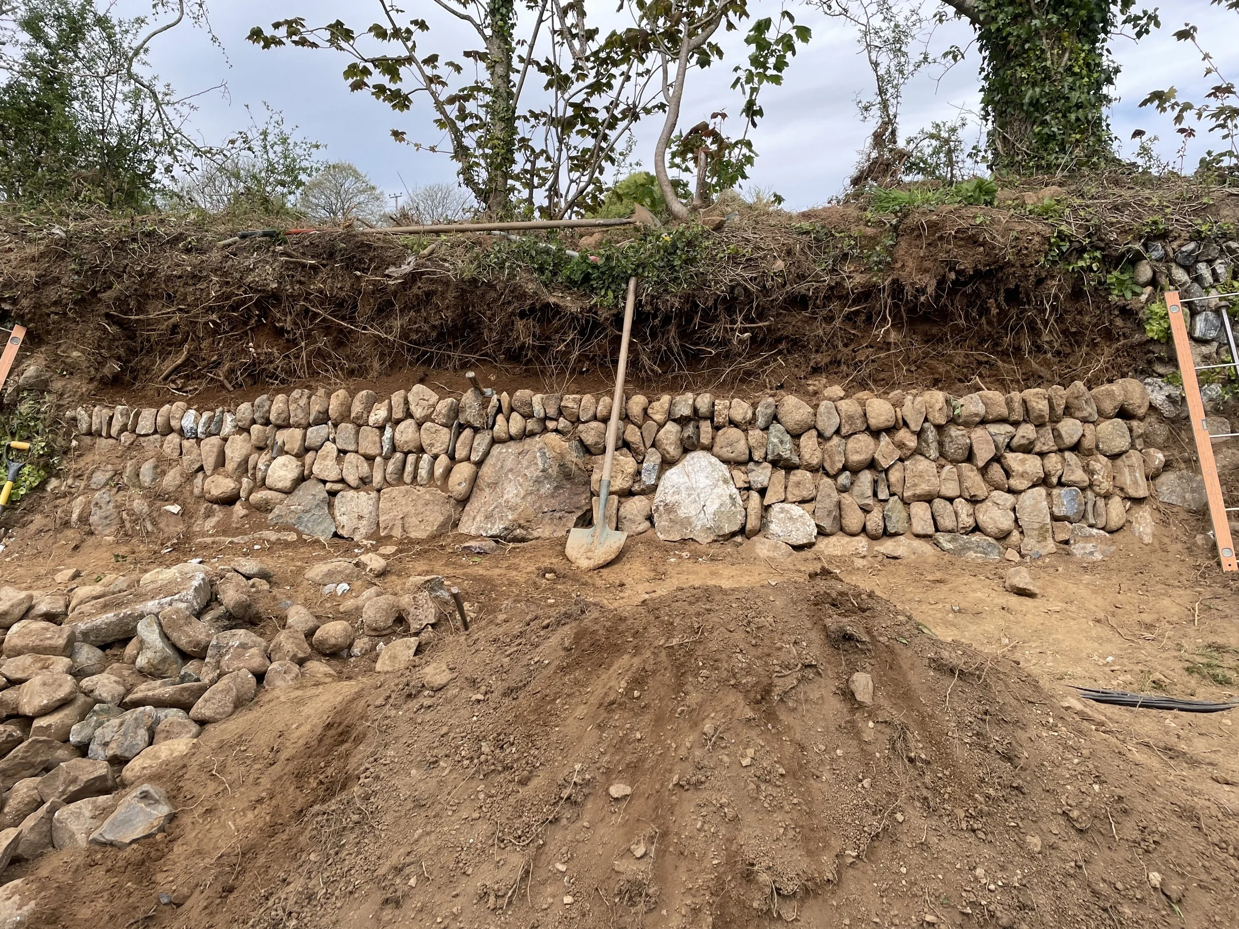 A construction or landscaping site with a stone retaining wall at the bottom, exposed soil, and some gardening tools and equipment. There are trees and cloudy sky in the background, with a shovel leaning against the dirt.