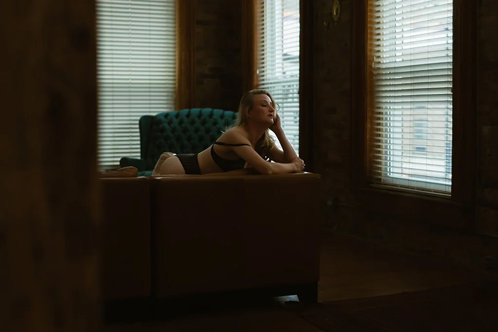 A woman lying on a couch in a dimly lit room with wooden walls, wearing black lingerie, gazing out the window blinds.