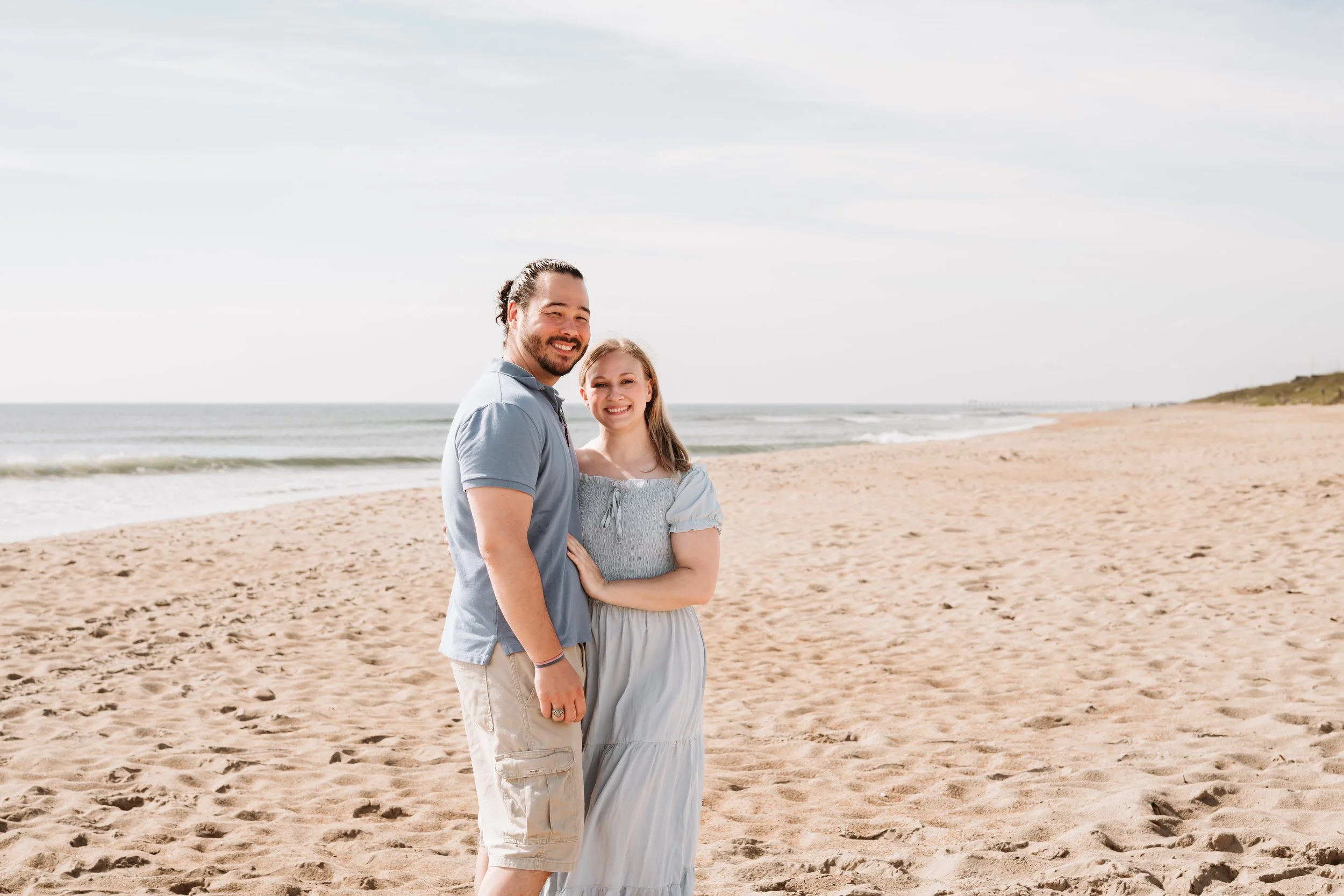 A smiling man and woman stand together on a sandy beach near the ocean, with a cloudy sky above.