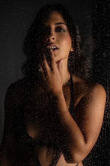 A woman with dark hair posing behind a glass window with raindrops, touching her face with one hand, looking at the camera through the rain.