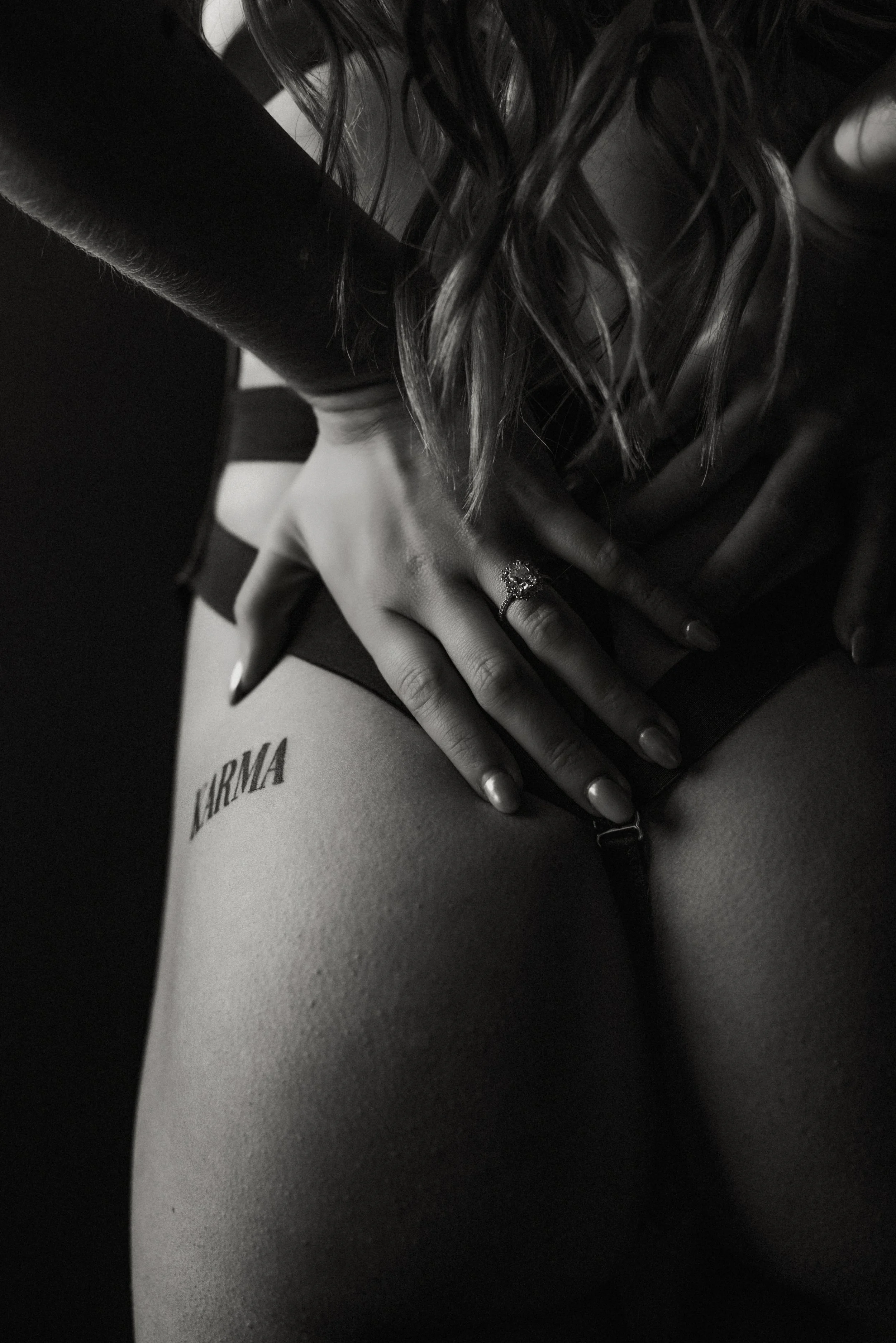 Close-up black and white photo of a woman’s hand resting on her lap, with a ring on her finger. She is wearing dark clothing, and her long hair is partially visible. The background includes a paper with partially visible text and a black object.