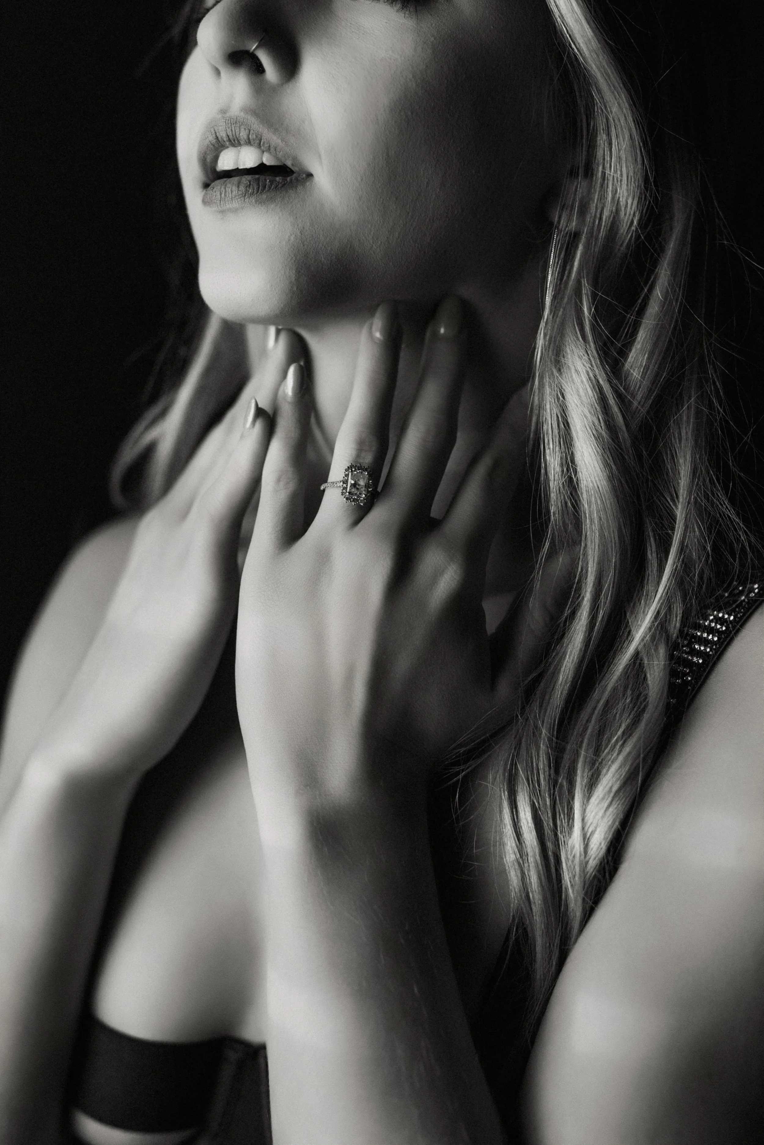 A black-and-white close-up photo of a woman's face and upper body. She has long, wavy hair and is wearing a ring on her finger. Her lips are slightly parted, and she has her hand gently touching her neck and chest.