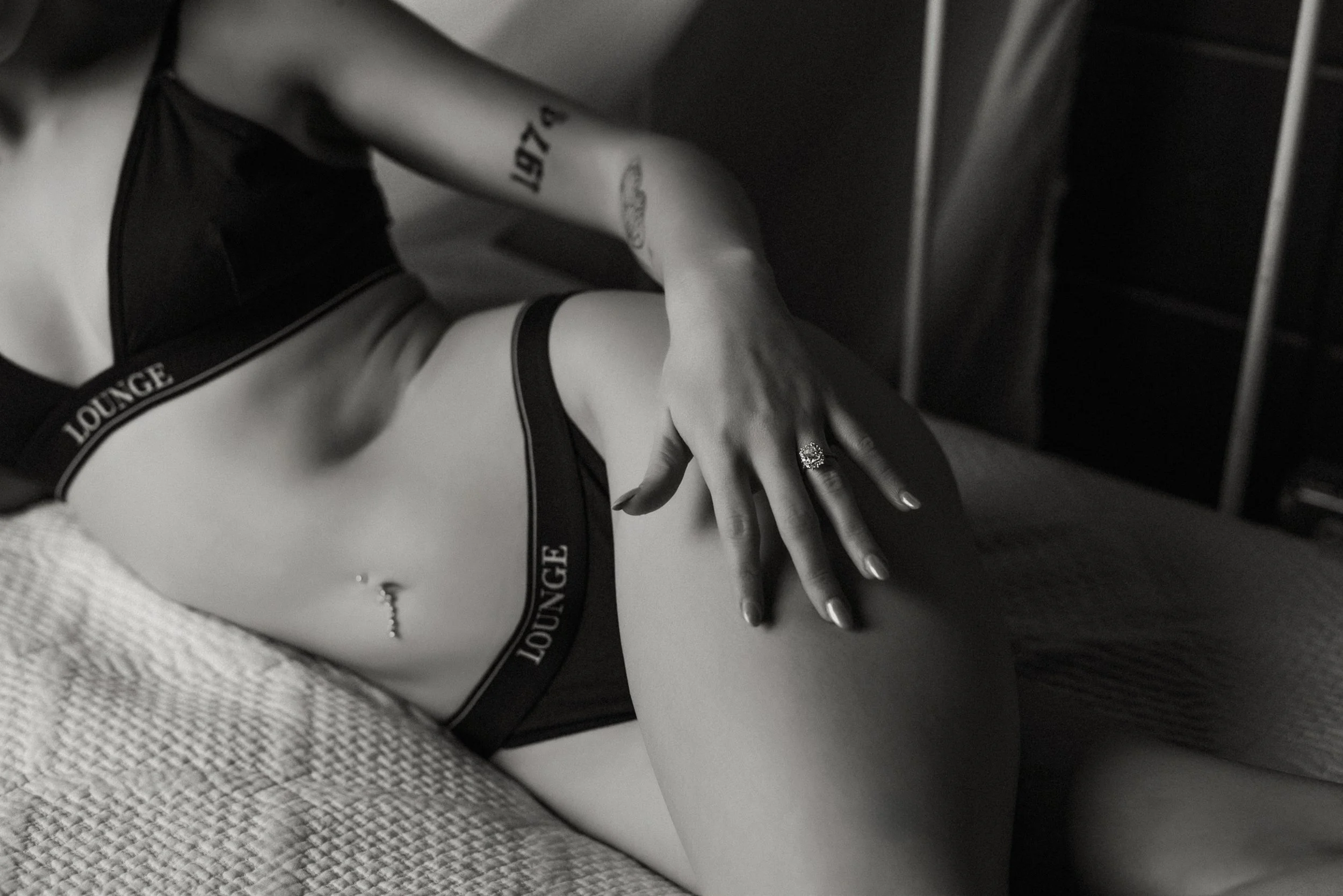 Black-and-white photo of a person's hand resting on their thigh, wearing a ring on their finger and a ring on their wrist. The person is dressed in minimal clothing, and some text is visible on their arm and underwear.