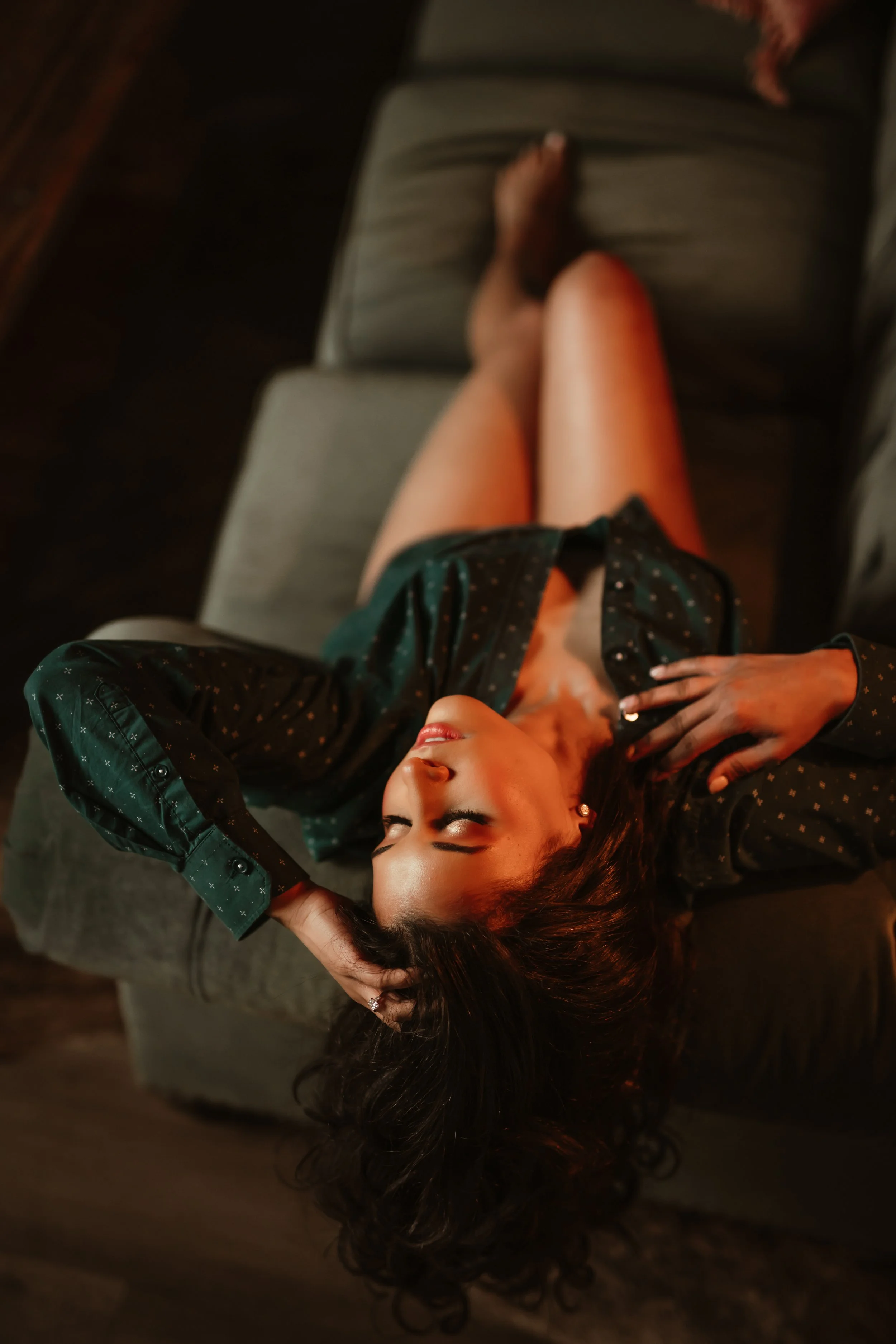 A woman is lying on her back on a sofa, with her eyes closed, one hand resting on her forehead and the other on her chest, wearing a dark polka dot shirt and shorts with a pattern, in a relaxed pose.