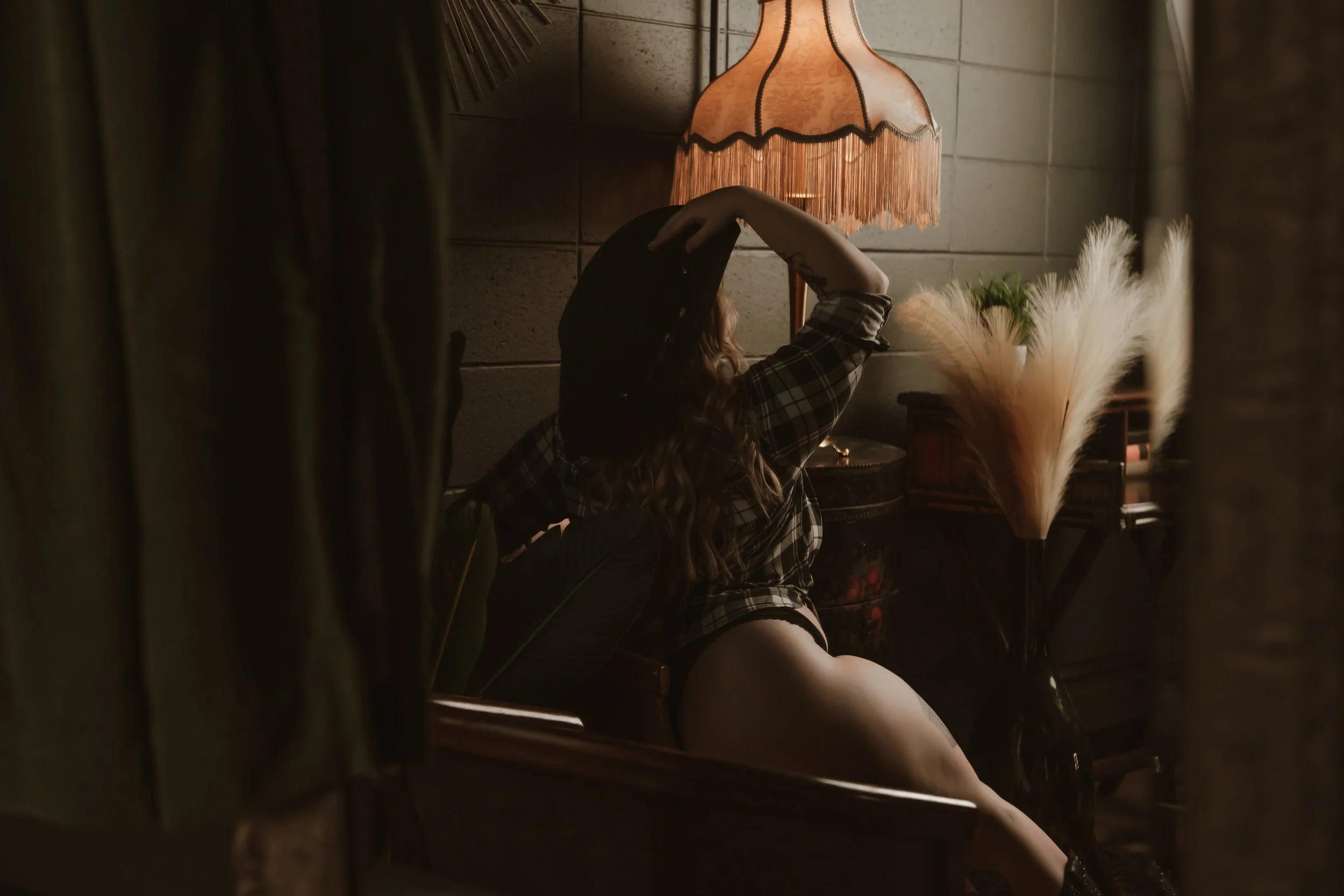 A woman with long wavy hair sitting on a piano bench in a dimly lit room, posing with one arm raised above her head, near a vintage lamp with a fringed shade, and decorative plants.