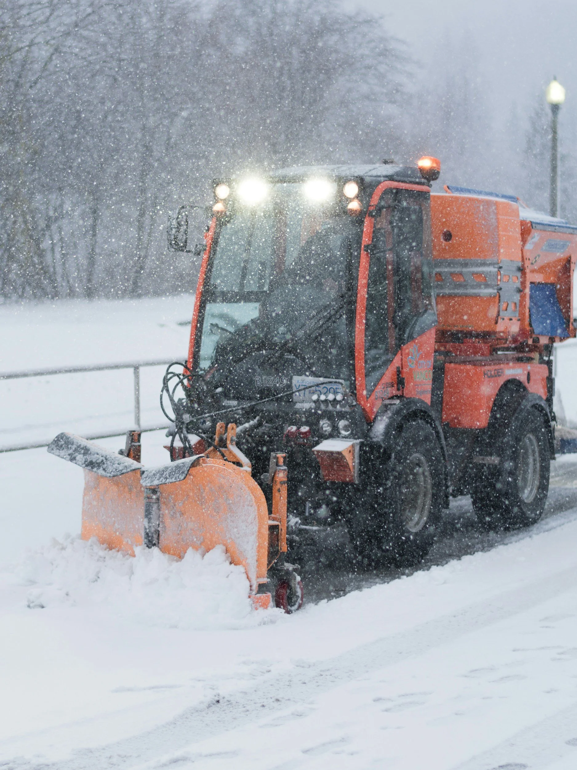 A snow plow clearing snow from a roadway during a snowstorm, with snow falling heavily and small snow piles on the ground.