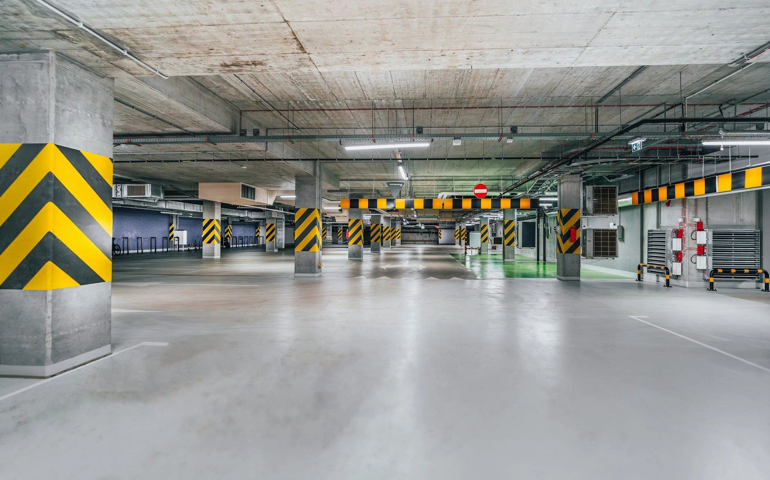 Empty underground parking garage with concrete columns painted with black and yellow stripes, ventilation units, bright ceiling lights, and emergency equipment.