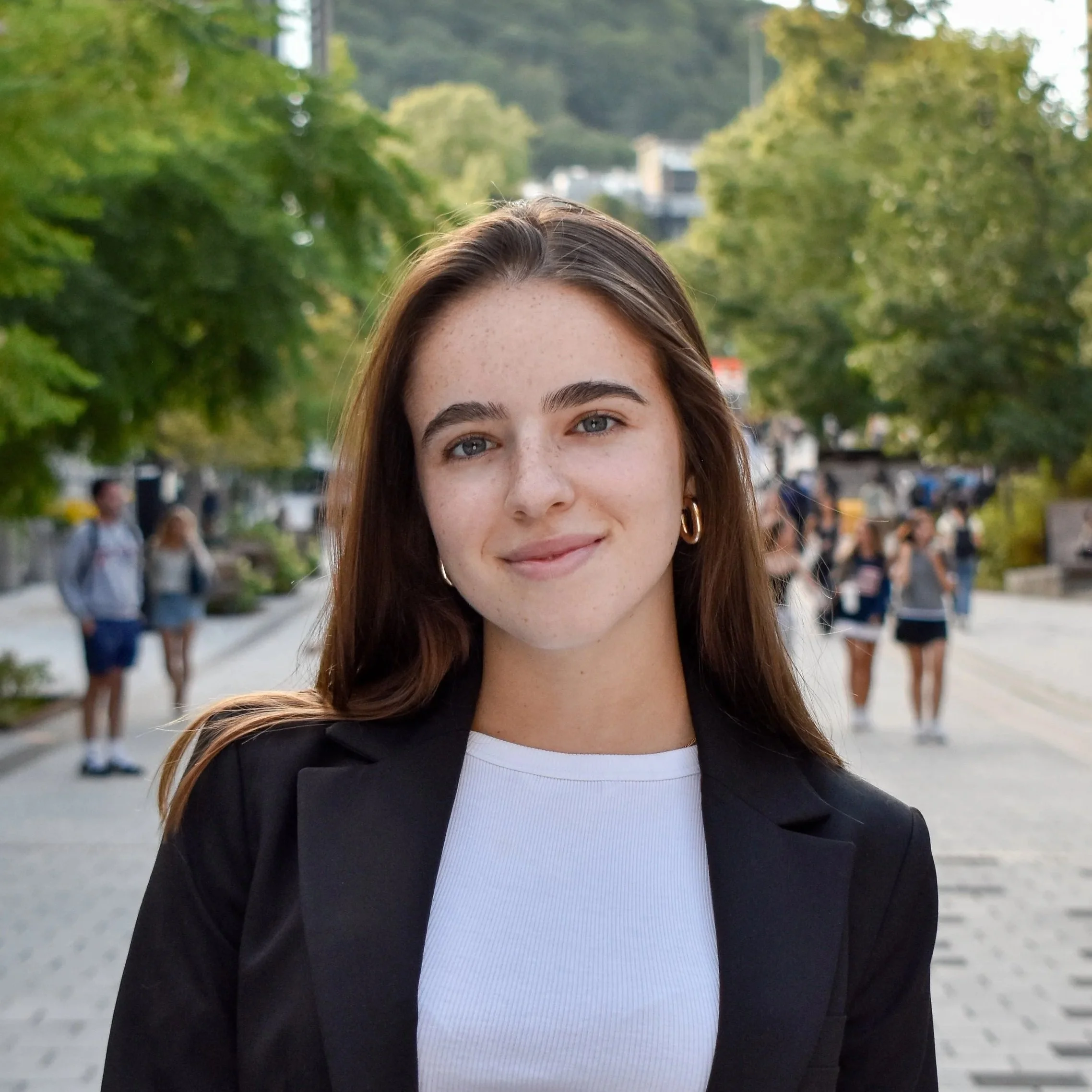 A young woman with long brown hair, blue eyes, and a nose ring, smiling outdoors during daytime, wearing a black blazer and white top, with trees and people walking in the background.