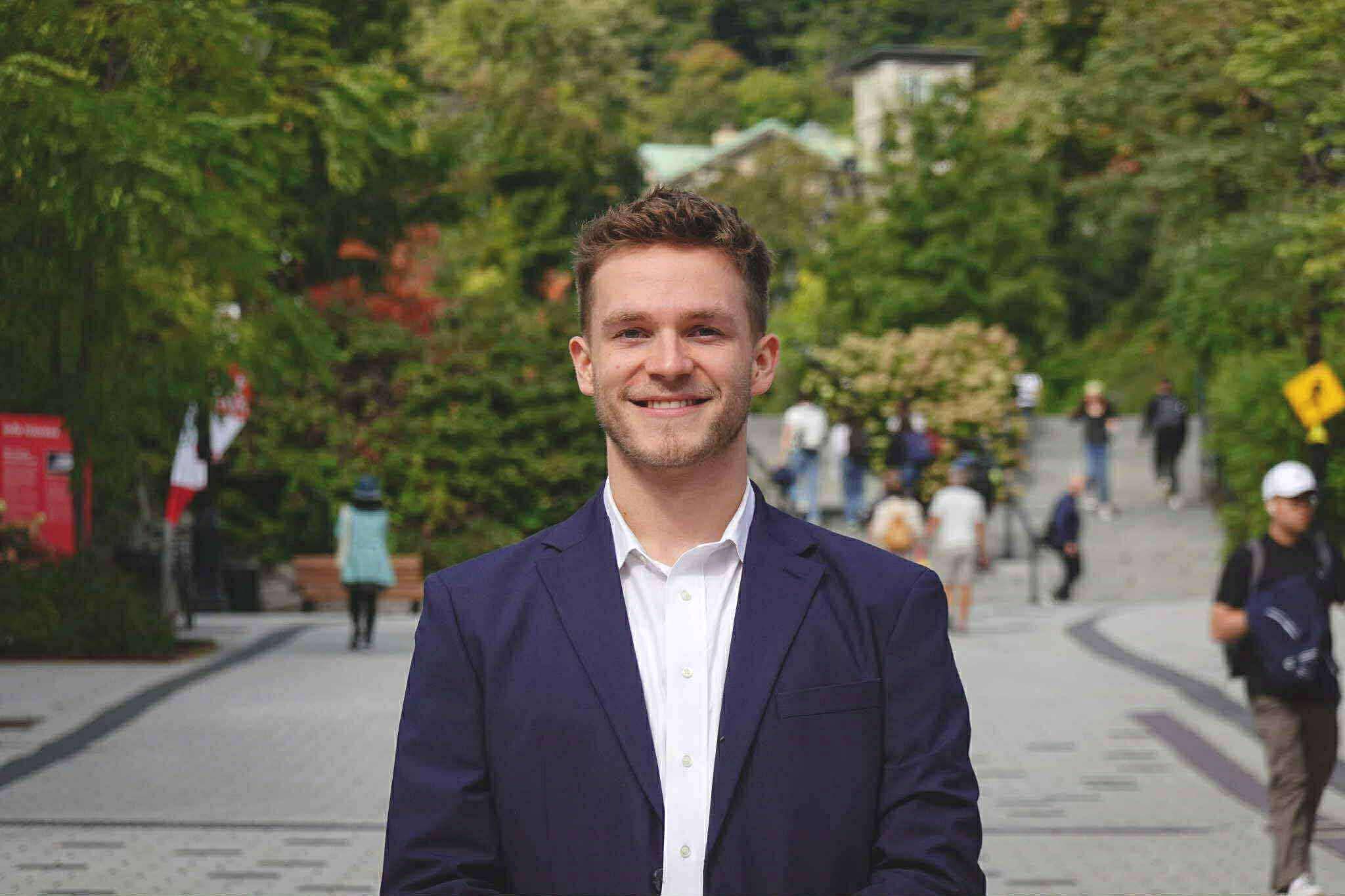 A smiling man in a dark blue blazer and white shirt standing outside in a park-like area with people walking in the background and greenery surrounds.