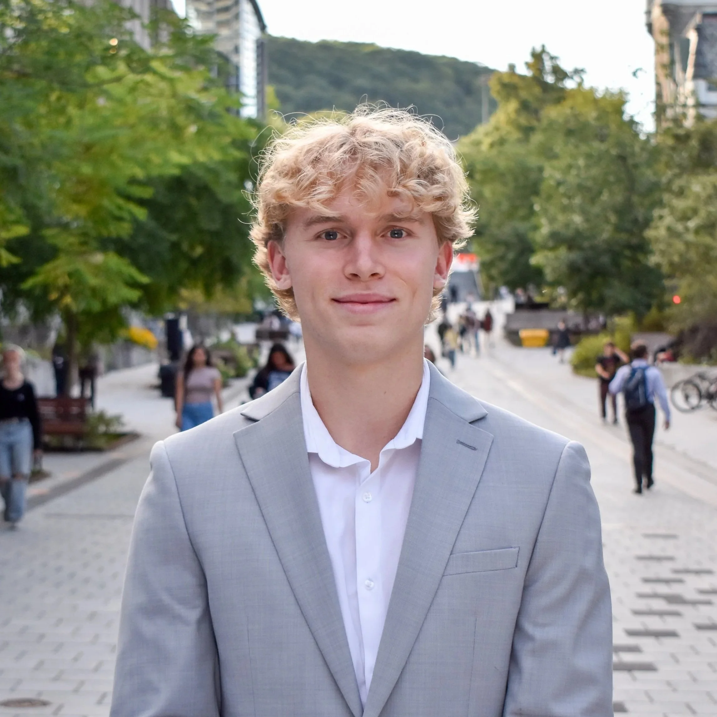 A young man in a light gray suit and white shirt standing outdoors on a city street with trees and buildings in the background, smiling at the camera.