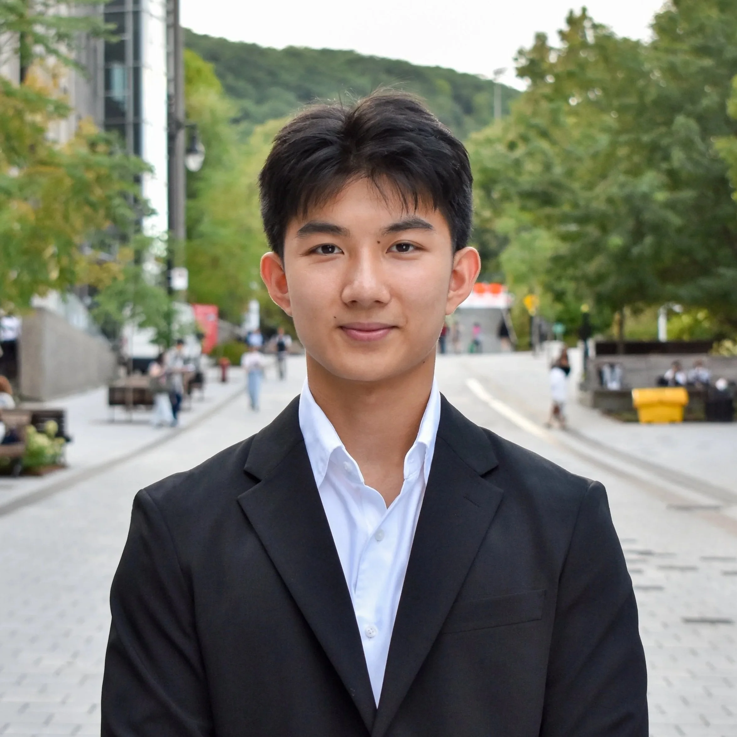 A young man in a black suit and white shirt standing outdoors on a city street with trees, benches, and people in the background.