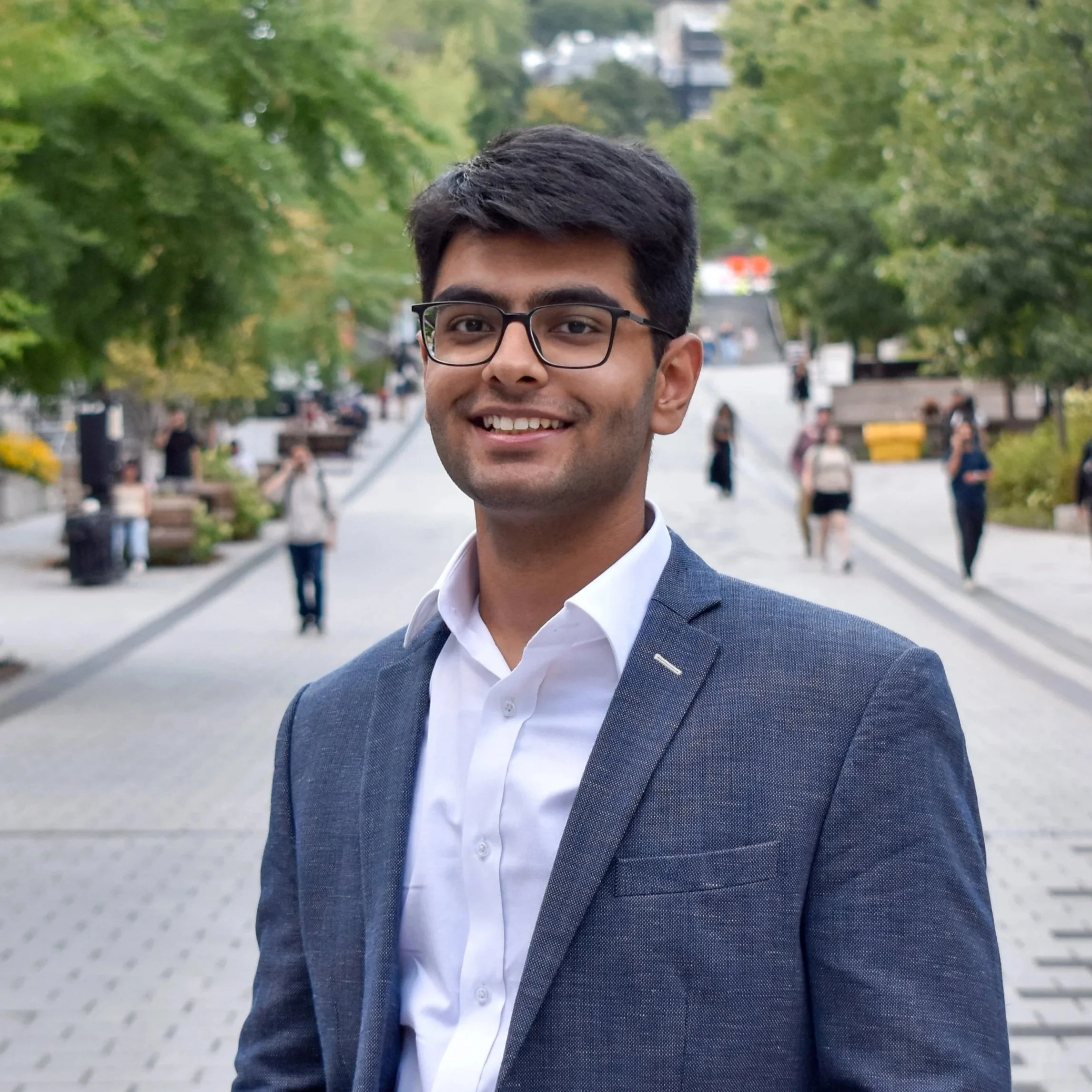 A young man in a gray blazer and white shirt stands outdoors on a city street with trees and pedestrians in the background.