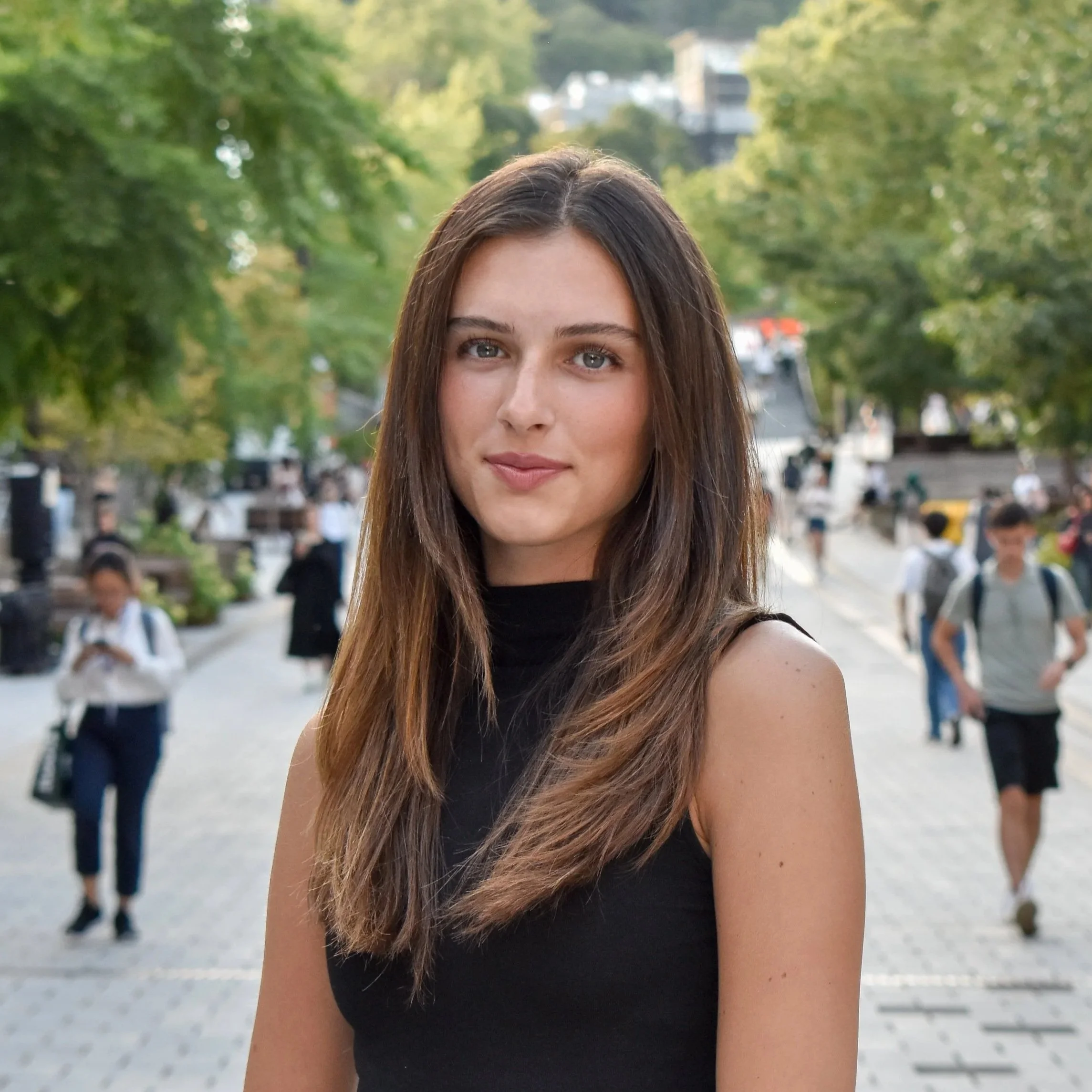 A young woman with long brown hair and blue eyes wearing a black sleeveless top standing outdoors on a city street with people walking and green trees in the background.