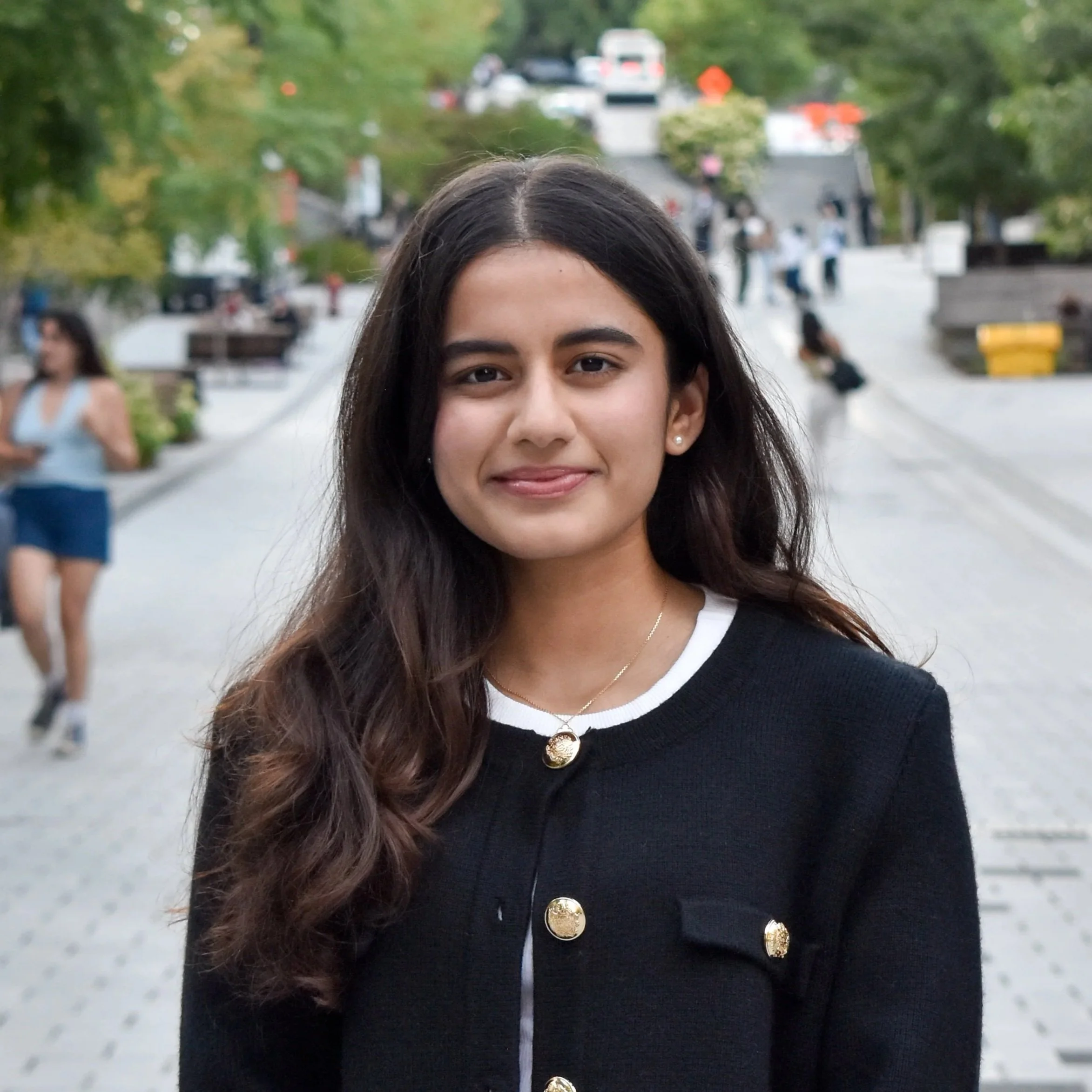 A young woman with long dark hair and a black cardigan with gold buttons, smiling outdoors in a park.