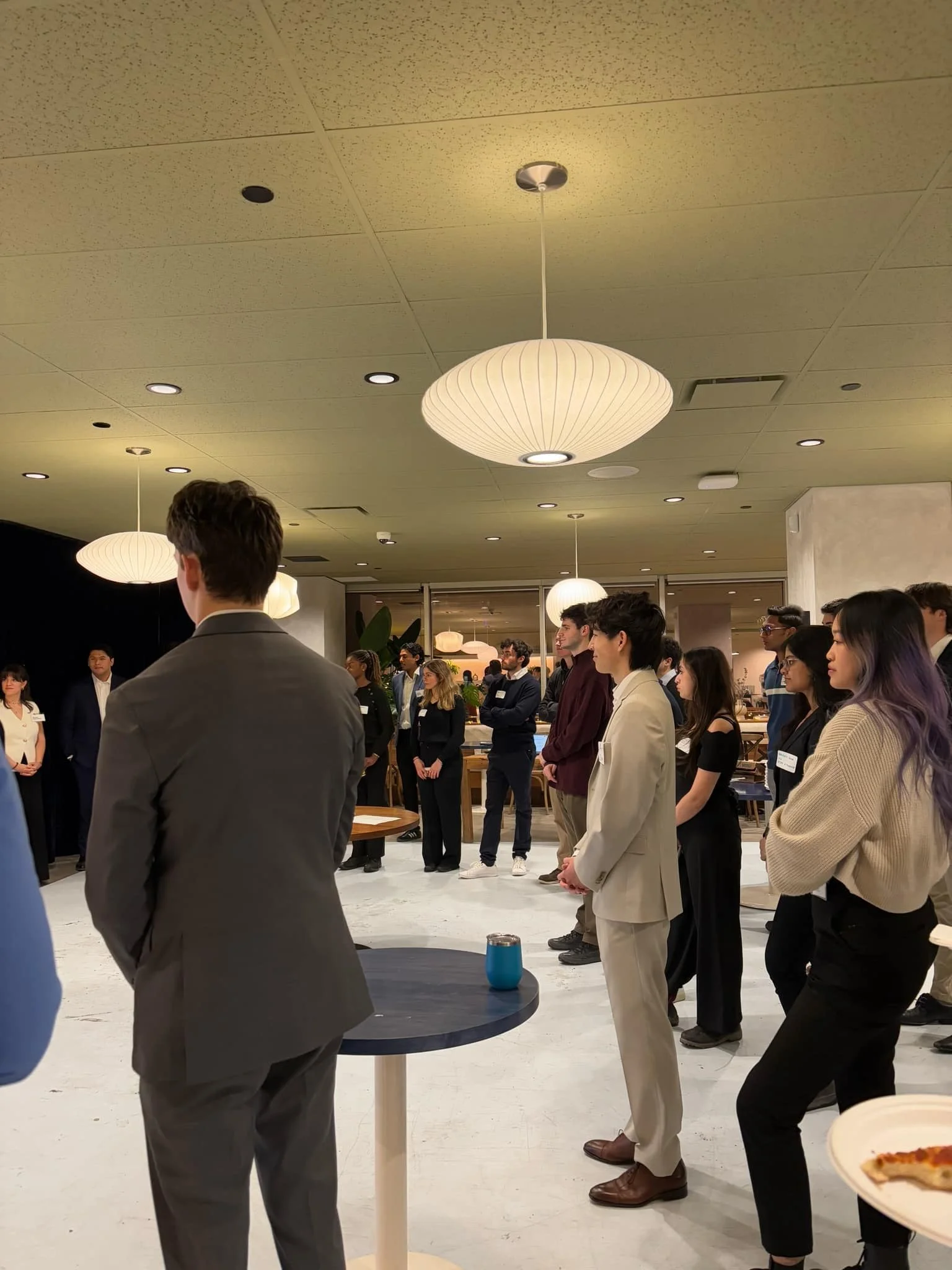 A group of diverse young professionals attending a networking event or conference indoors, standing and listening, with some holding drinks.