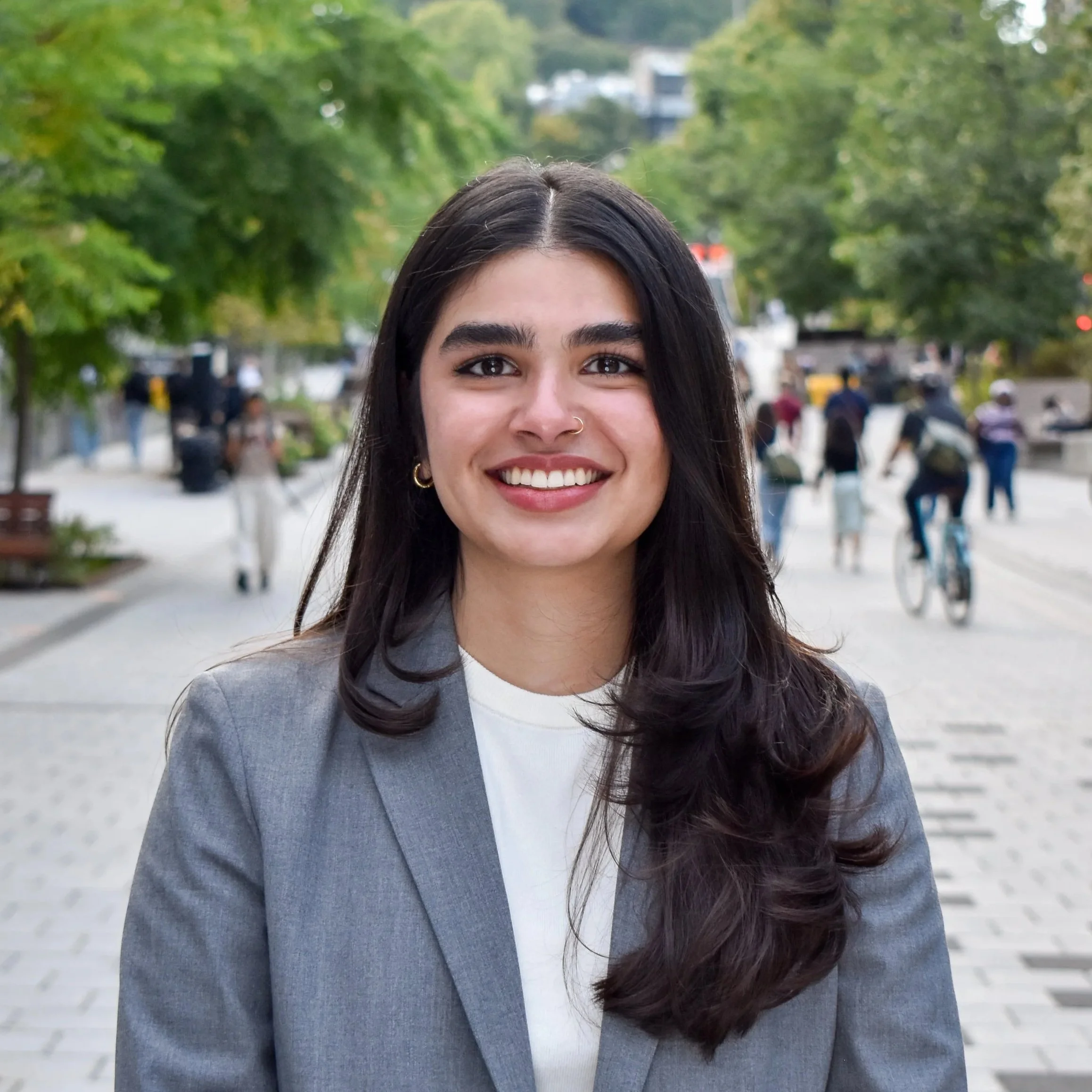 Smiling woman with dark hair, wearing a gray blazer and white shirt, standing on a city street with trees and people in the background.