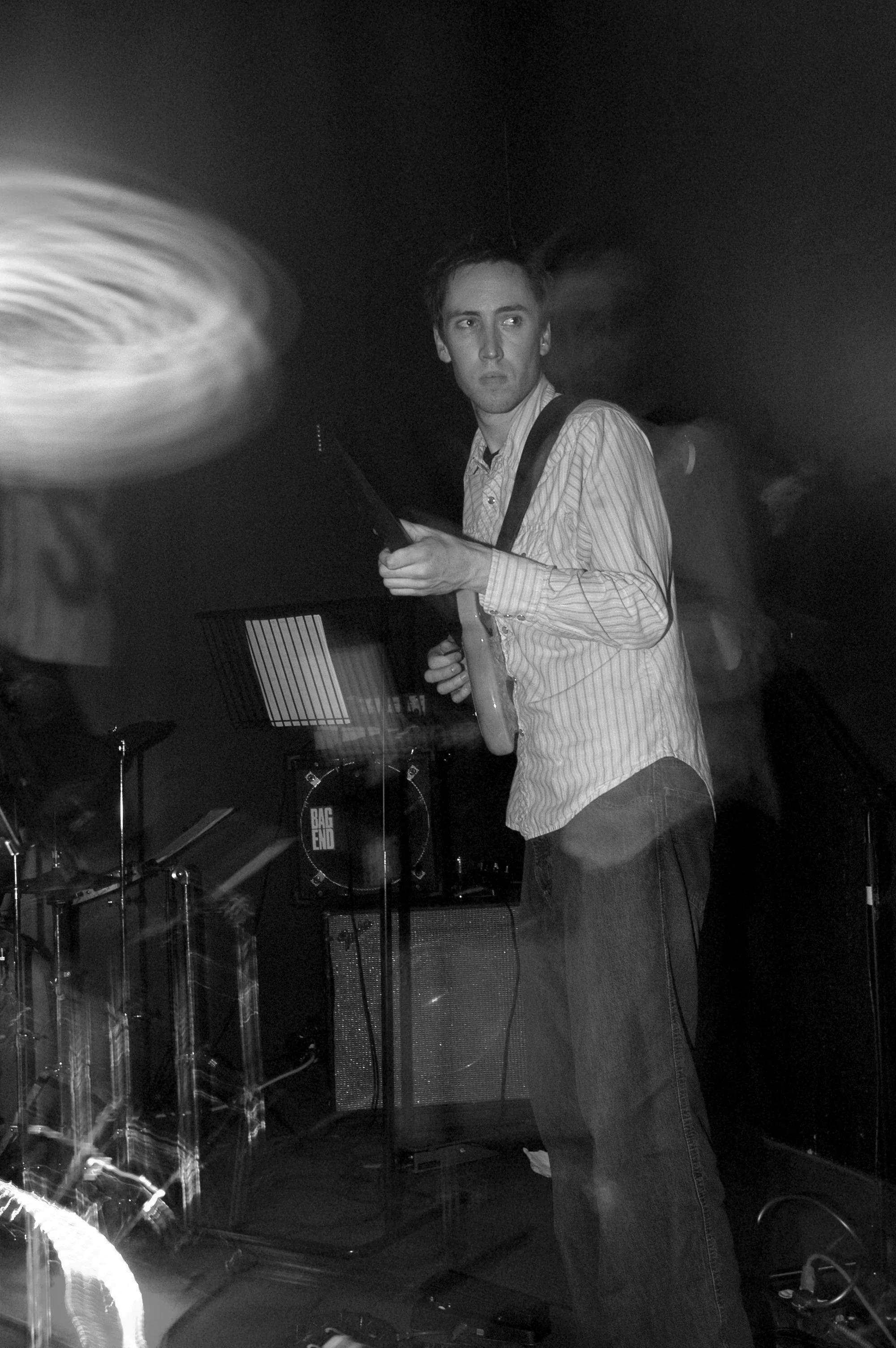 A young man with short hair wearing a striped shirt stands in a music studio, holding a guitar and looking to his right. Musical equipment, including drum set and amplifiers, are visible in the background, and the scene is in black and white.
