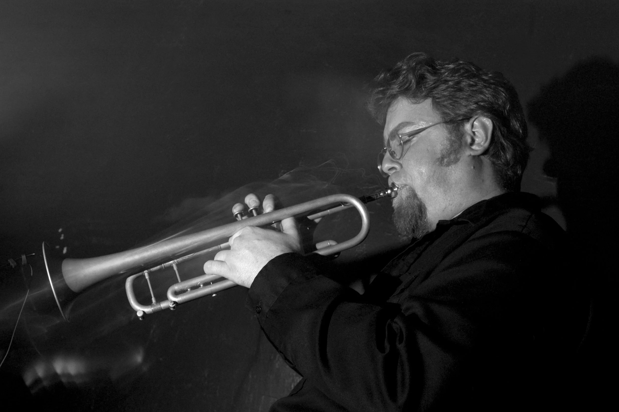 A man with glasses and facial hair playing a trumpet in a dark setting.