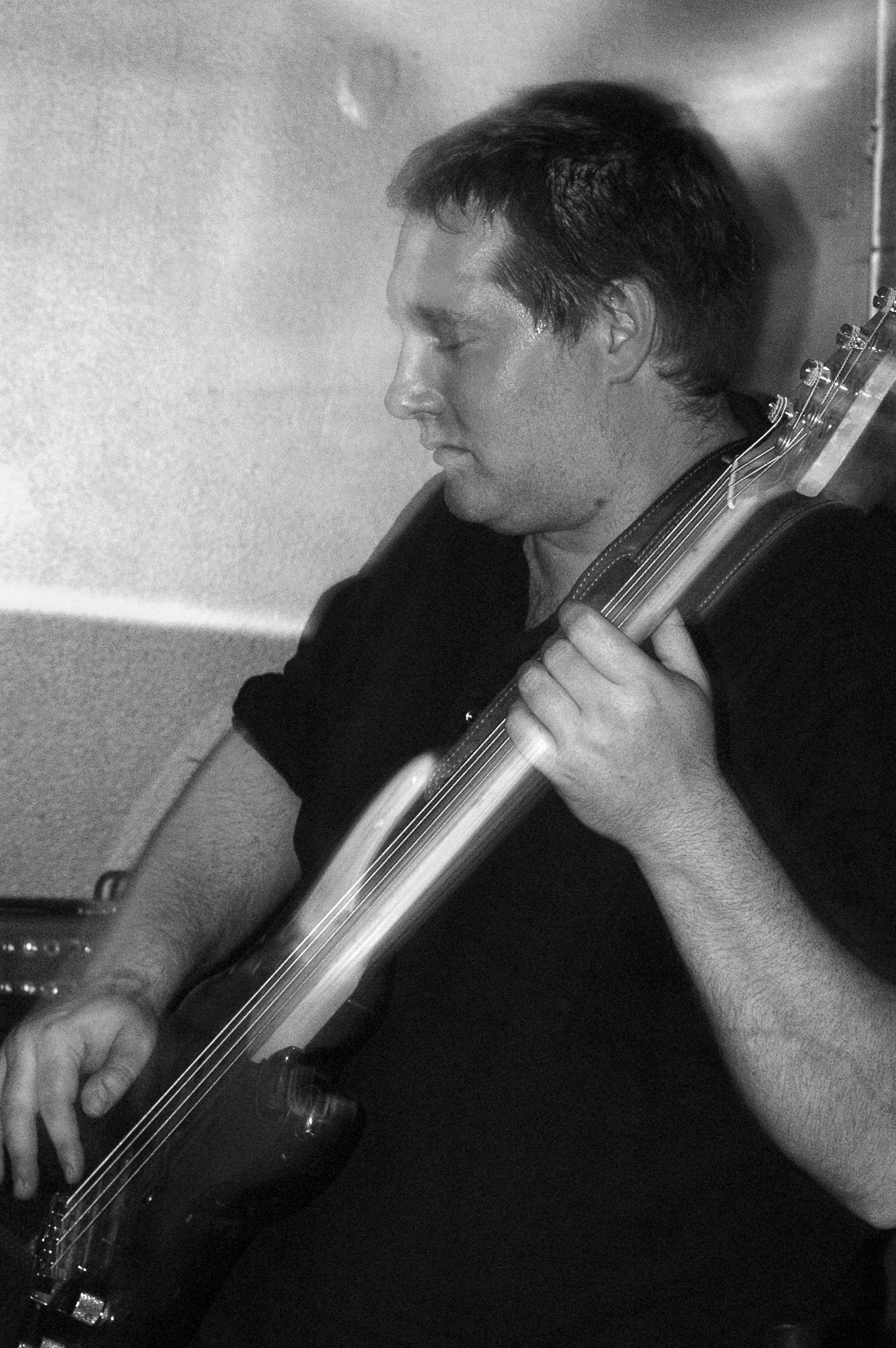Black and white photo of a man with dark hair playing an electric guitar, with his eyes closed, in front of a plain wall.