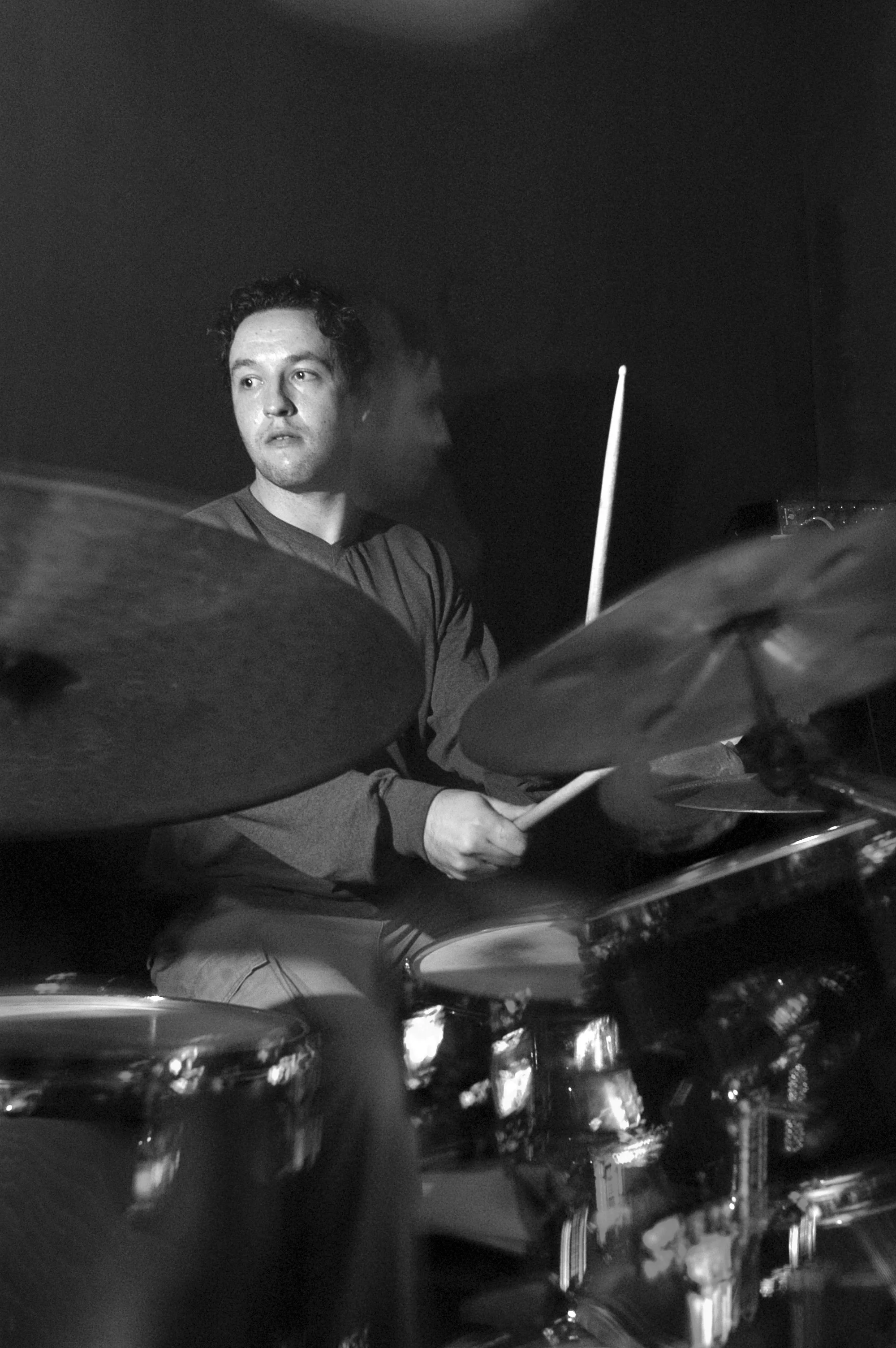 A black and white photo of a man playing the drums, with his face partially visible, surrounded by a row of car headlights.
