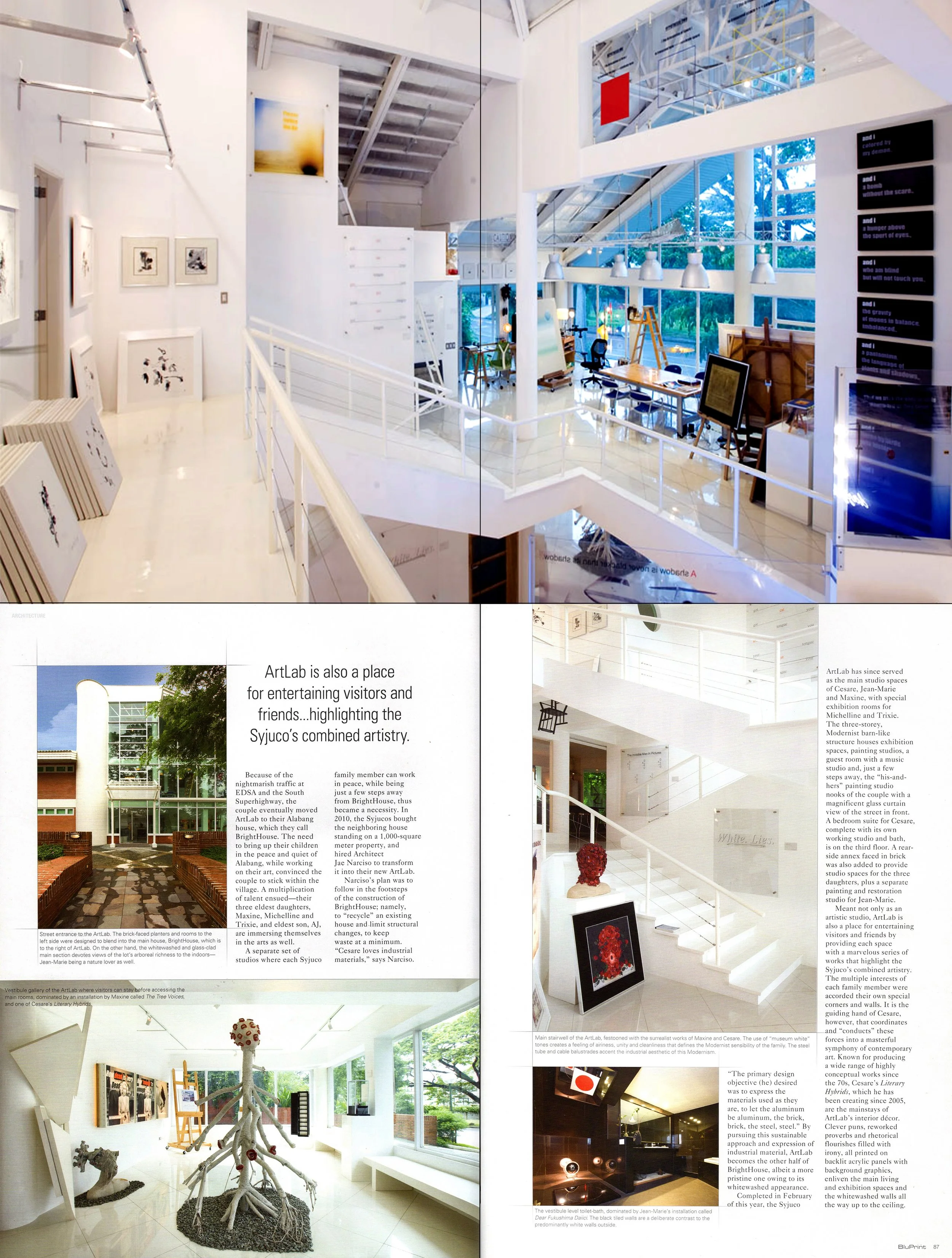 Interior view of an art gallery with white walls, artwork on display, and modern lighting fixtures. There is a staircase, large windows showing outside greenery, and various art pieces including framed artworks, sculptures, and digital displays.