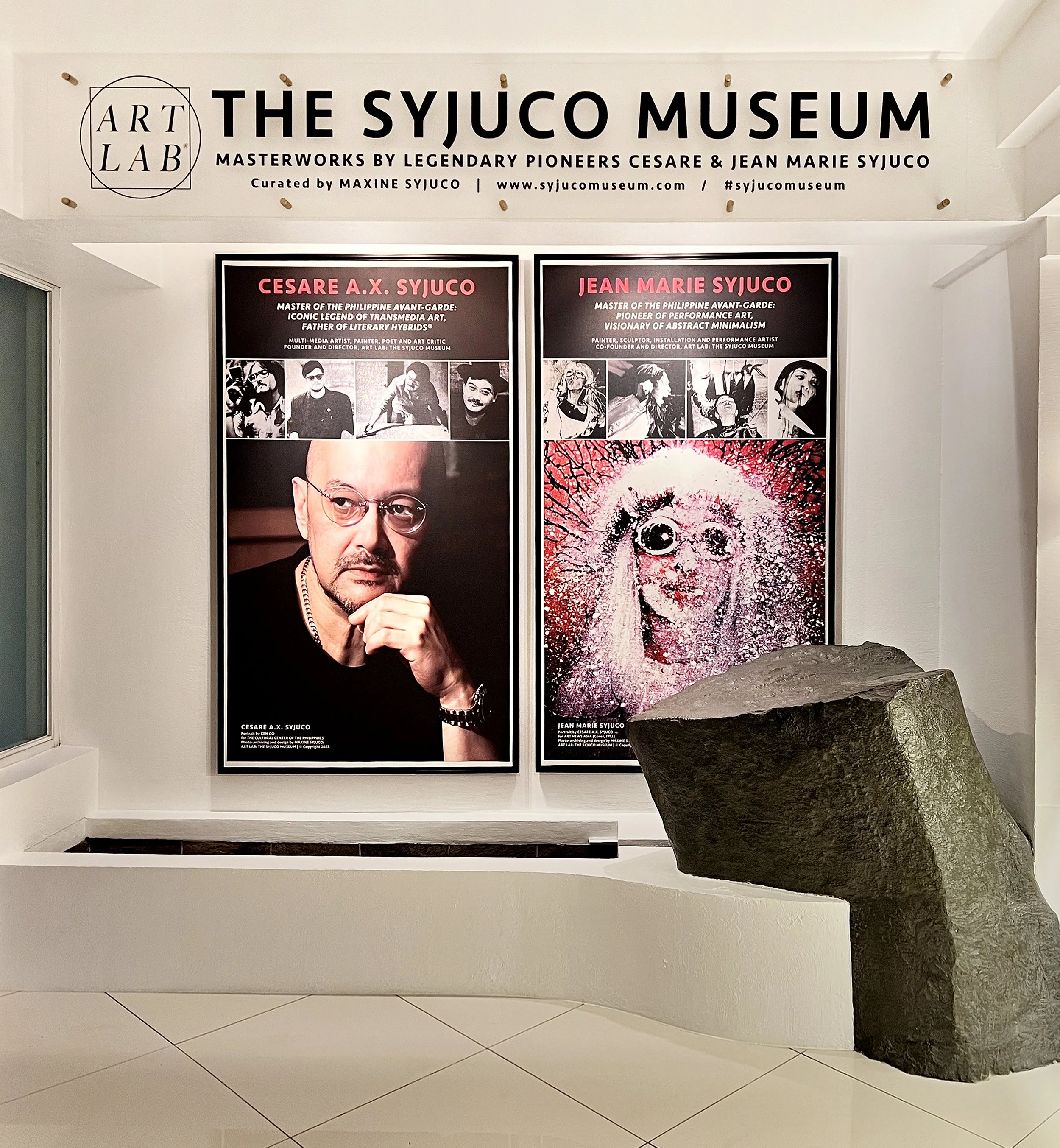 Preview image of Art Lab The Syjuco Museum featuring portraits of Cesare A.X. Syjuco and Jean Marie Syjuco with descriptive text, displayed on a white wall with a large dark rock sculpture in the foreground. Curated by museum director Maxine Syjuco