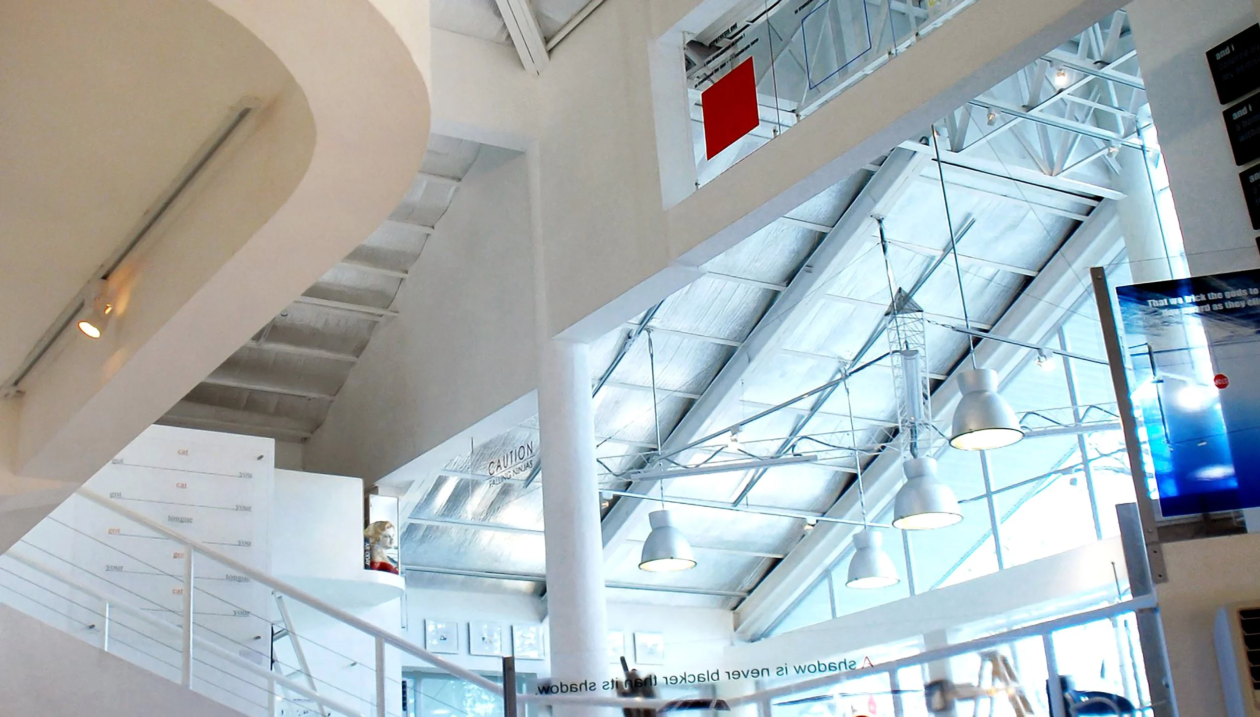 Interior of a modern building with multiple levels, featuring white railings, large windows, and hanging lights, with industrial-style ceiling beams and a digital display screen.