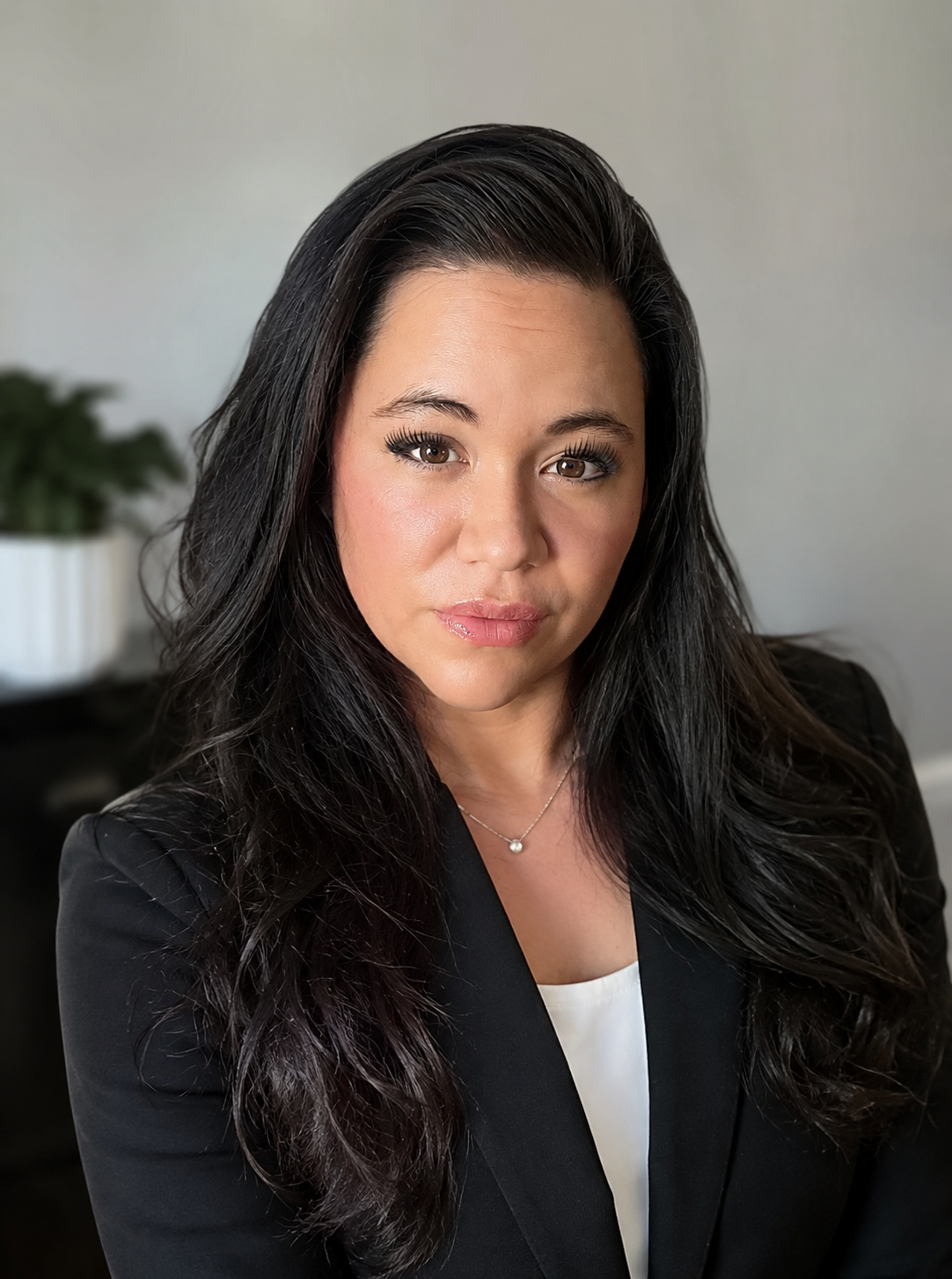 Professional woman with long dark hair wearing a black blazer and white top, posing indoors with a neutral background and a potted plant in the blurred background.