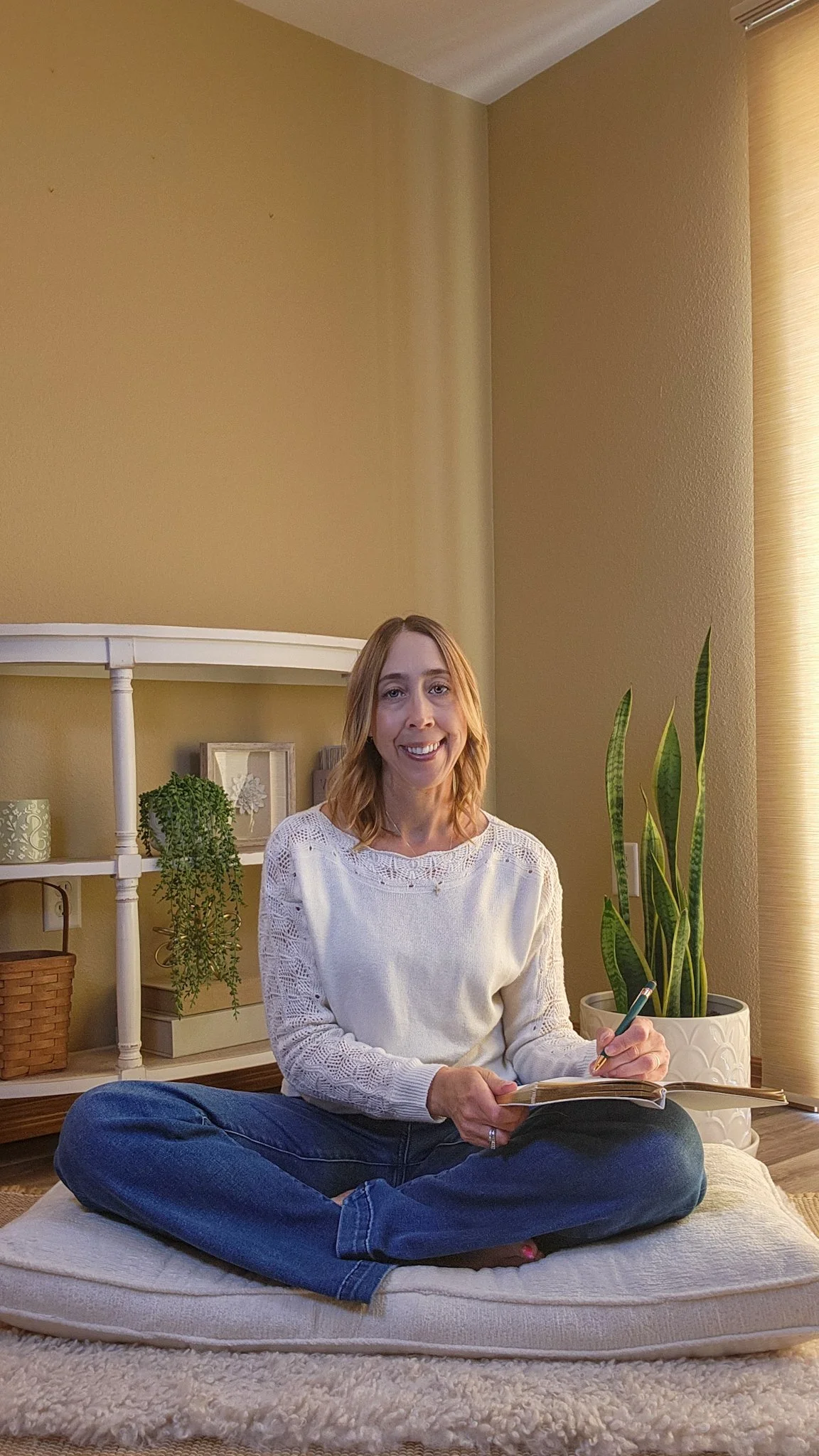 A woman sitting cross-legged on a cushion on the floor, smiling, holding a notebook and pen, in a cozy room with plants and bookshelves.