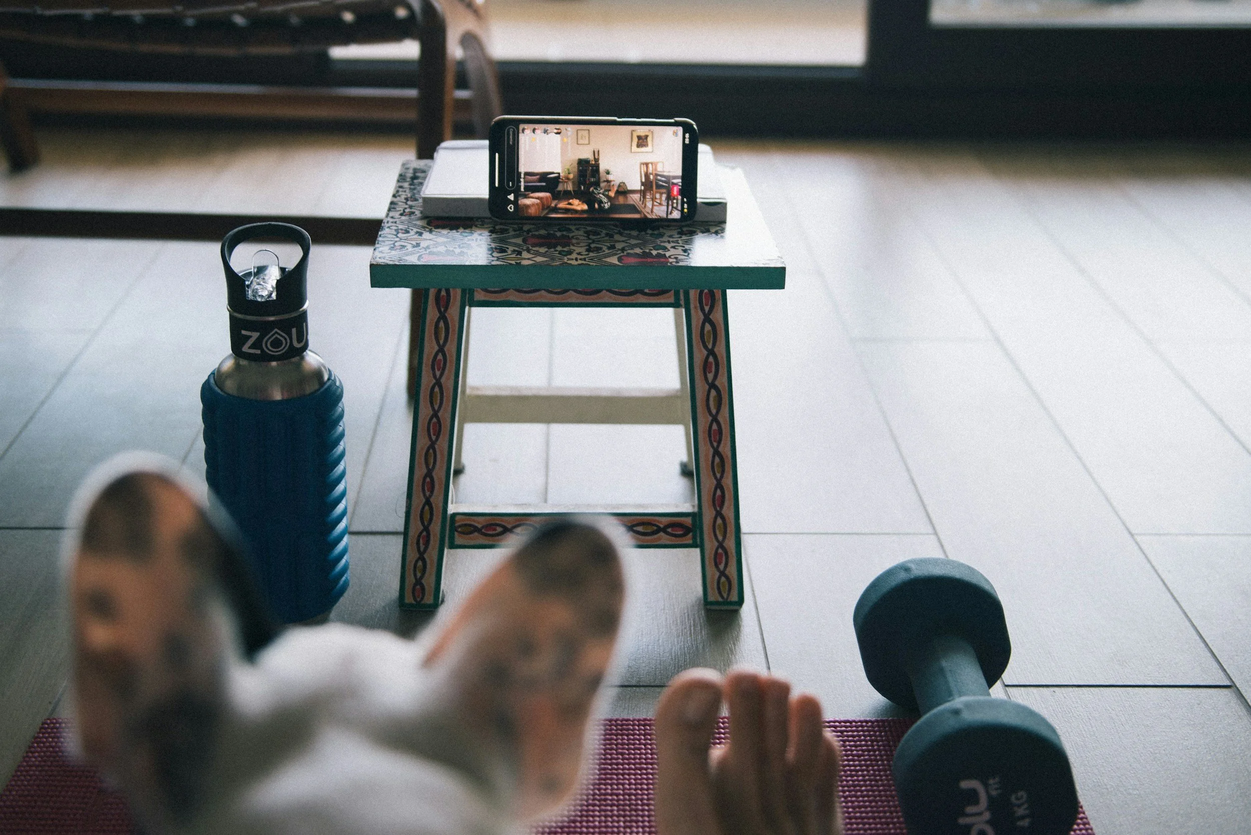 A person in white sneakers is lying on a purple yoga mat, taking a selfie with a smartphone. In the background, there are dumbbells, a small table, and a decorative outdoor tile table with a colorful pattern. The setting appears to be indoors near a large window or glass door showing an outdoor area. A water bottle with a black lid and a blue sleeve is on the floor to the left of the person.
