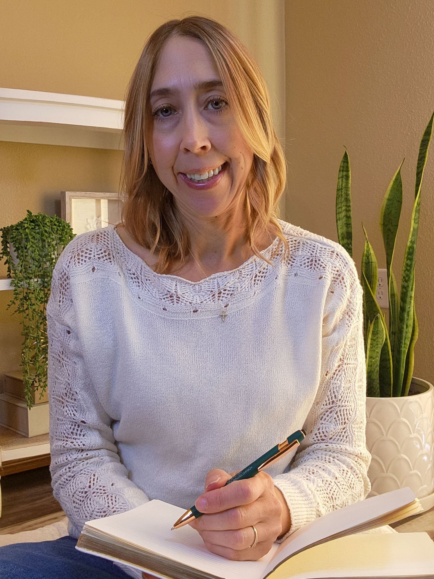 A woman with shoulder-length blonde hair, wearing a white lacy sweater, sitting at a table, holding a pen and an open notebook, with plants and a beige wall in the background.