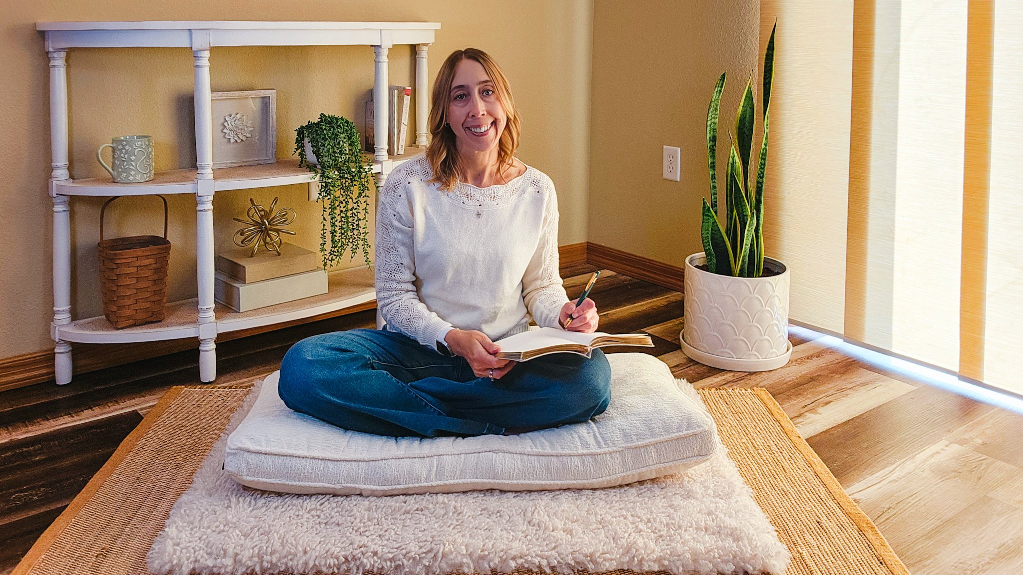 A woman sitting cross-legged on a floor cushion, smiling while holding a notebook and pen, in a warmly lit room with a plant and a bookcase behind her.