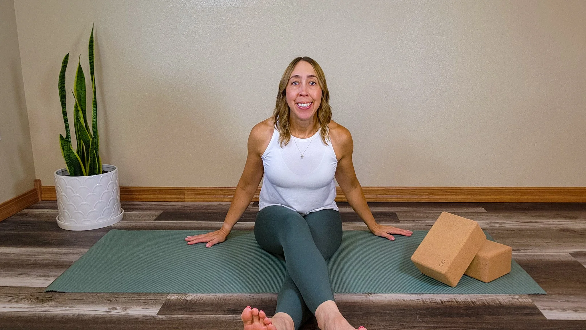 A woman practicing yoga indoors on a green yoga mat, sitting in a seated twist pose with a yoga block nearby, a large indoor plant in a white pot, and a plain beige wall.