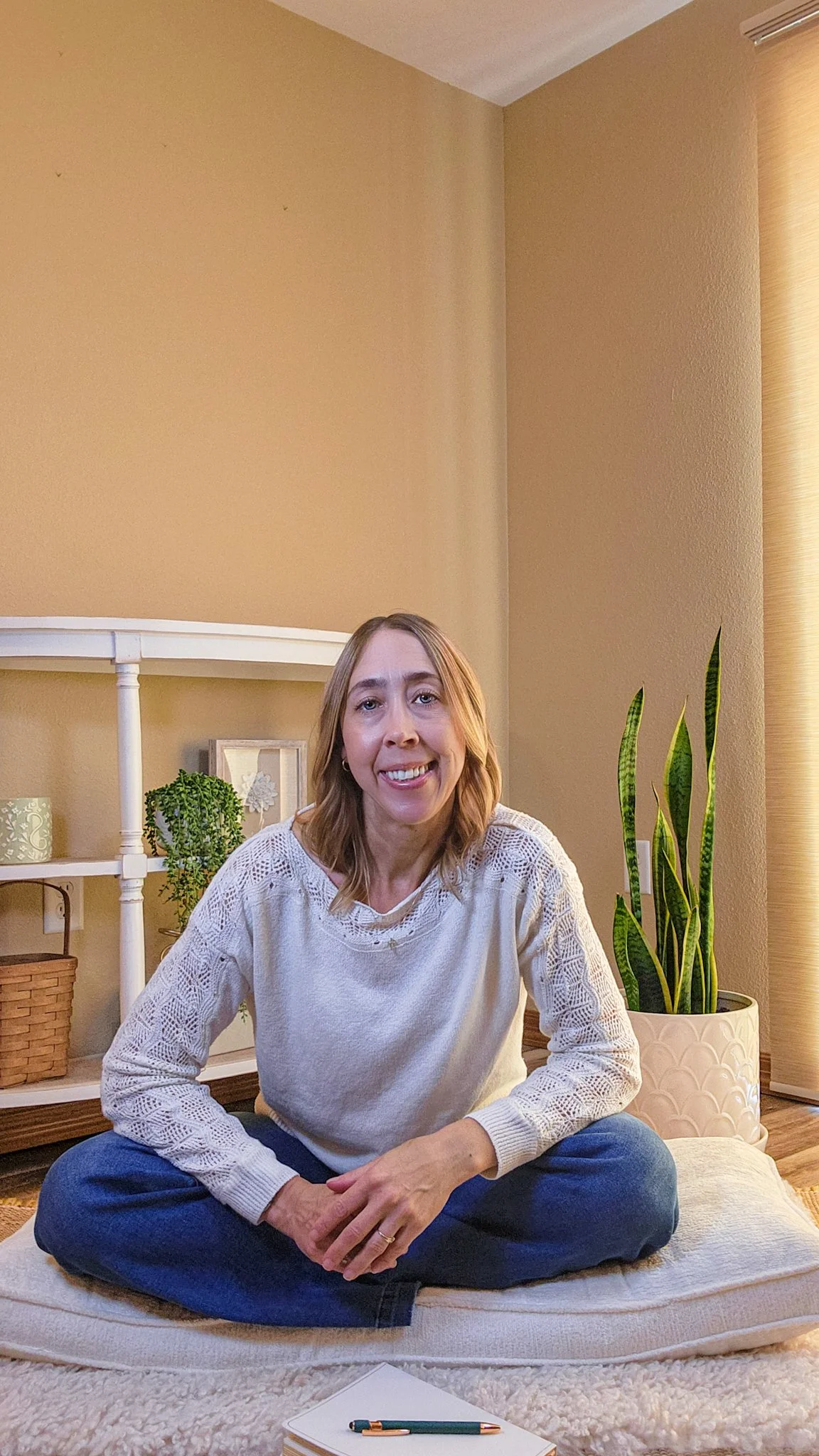 A woman with shoulder-length light brown hair, smiling, sitting cross-legged on a cushion on a cream-colored carpet in a cozy room with beige walls. There are houseplants, a white shelf, and decorative items in the background.