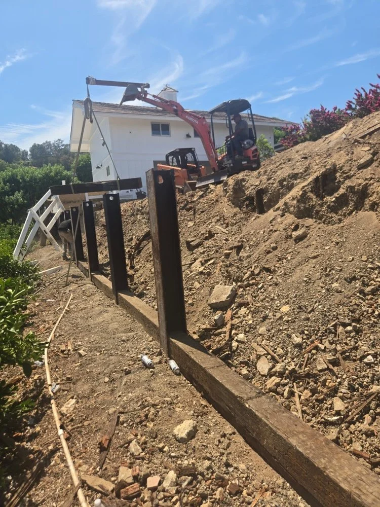 Hillside retaining wall installation with structural steel I-beams, excavation, and grading by Big City Construction