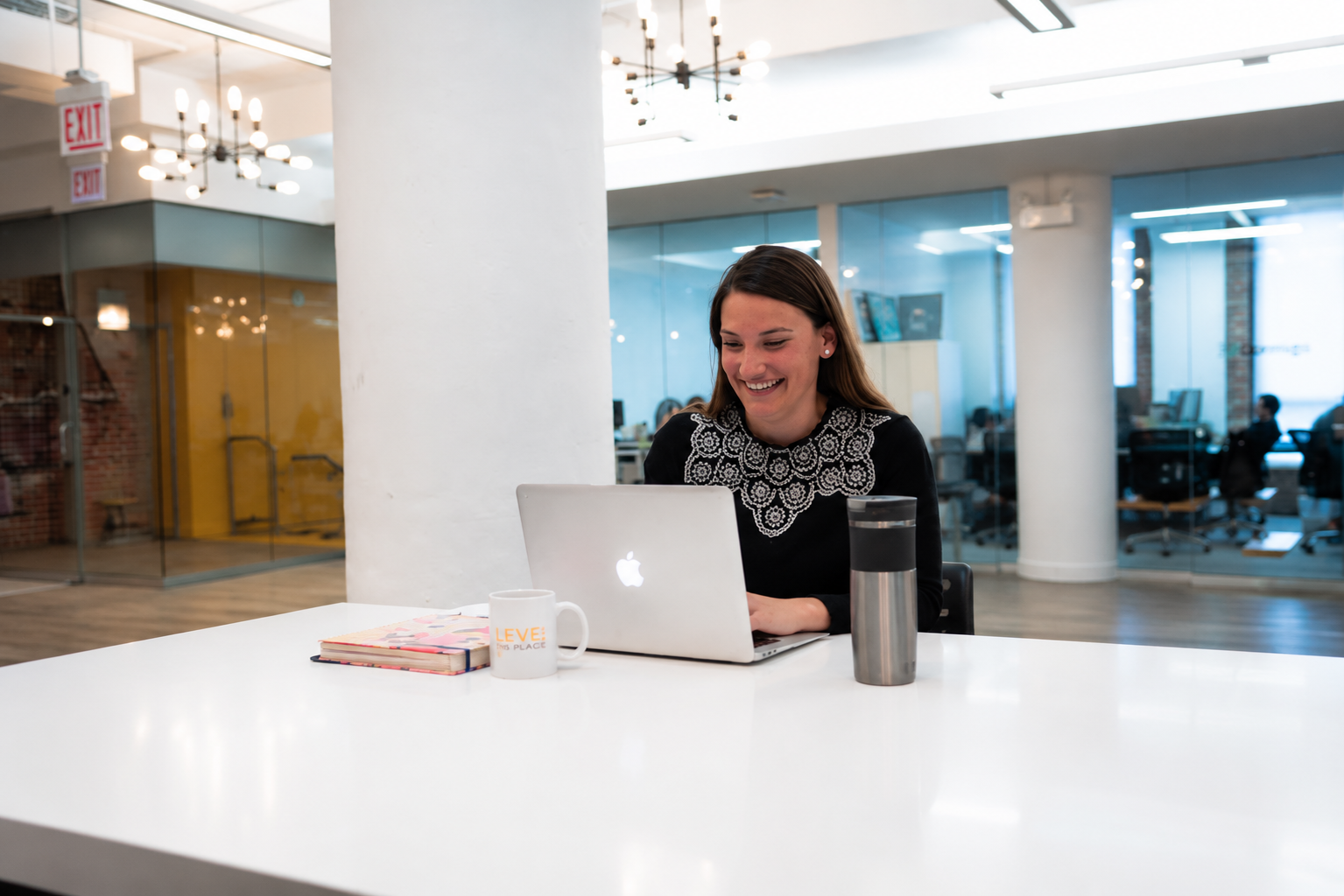 A woman with long brown hair, wearing a black top with lace detailing, sitting at a white table in a modern office space. She is smiling while working on a silver MacBook. On the table, there is a white coffee mug, a closed notebook, and a stainless steel water bottle. The background features glass walls, white columns, and other people working at desks.
