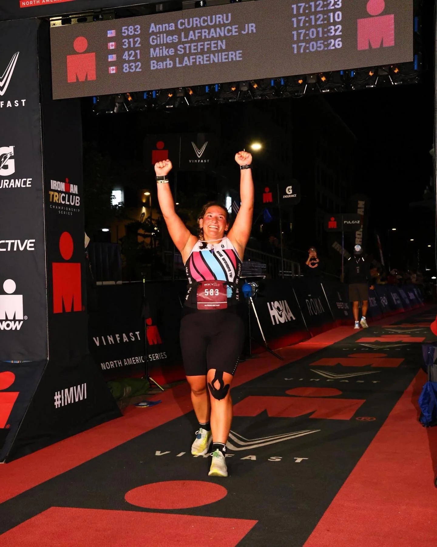 Female athlete finishing a race with her arms raised in celebration, crossing the finish line at night. She is smiling, wearing a black sports outfit and a race bib number 583. The background features a digital leaderboard displaying names and times of other competitors, sponsor banners, and event signs.