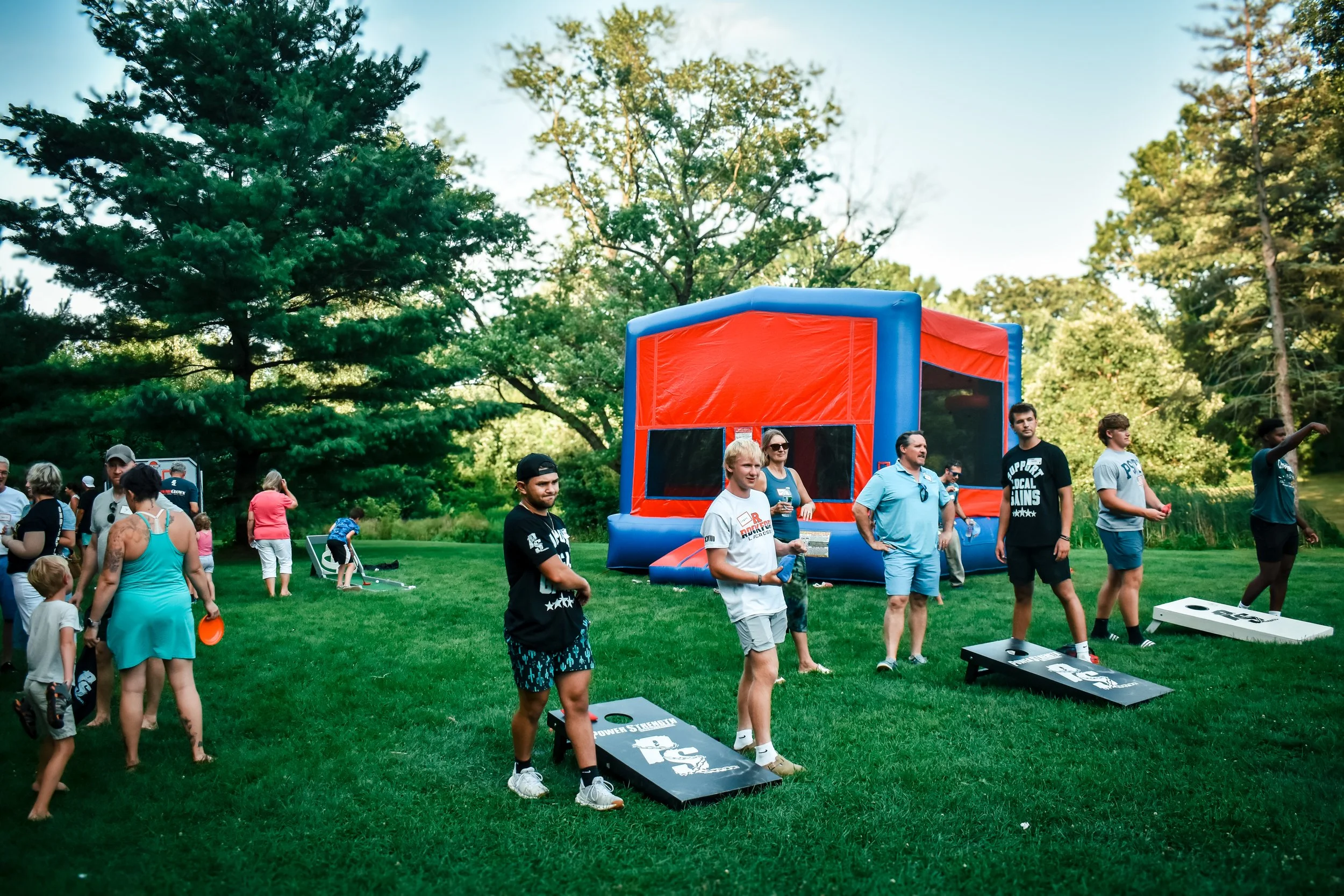 People gathered outdoors in a park for a community event with a large inflatable obstacle course, cornhole games, and many trees in the background.
