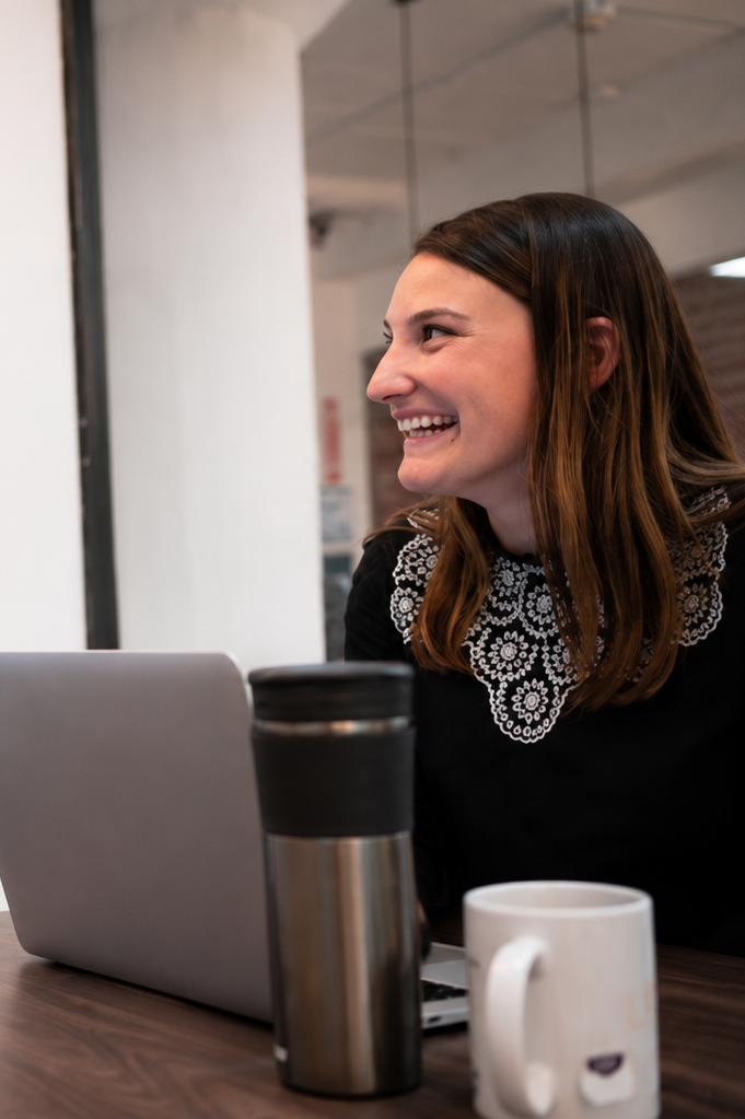 A young woman with brown hair and a black top with white embroidery, smiling and talking to someone off-camera, sitting at a table with a laptop, a stainless steel travel mug, and a white coffee mug.