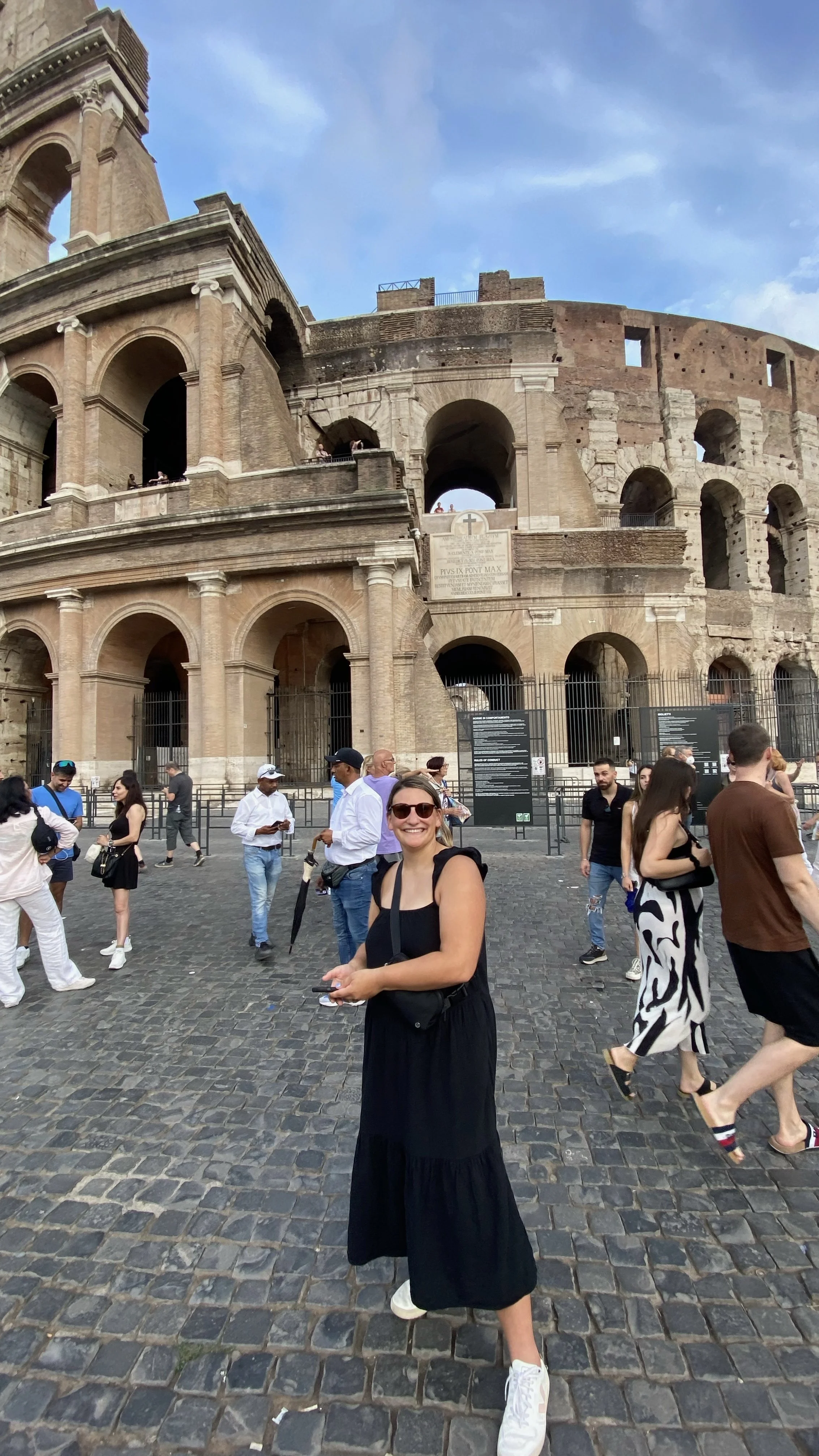 People standing in front of the Colosseum in Rome, Italy, with some tourists taking photos and smiling.