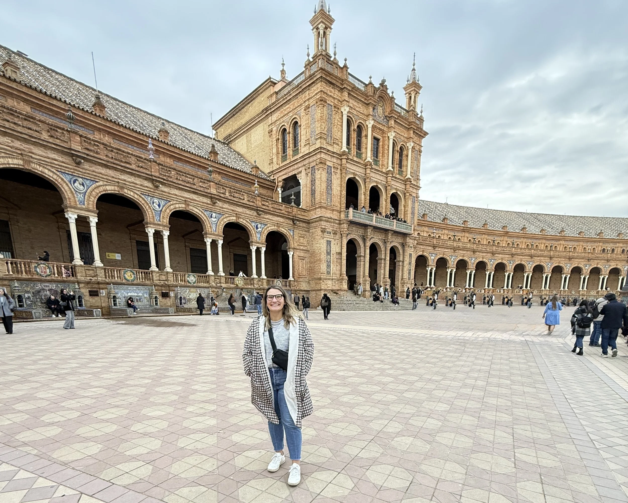 A young woman standing in front of the Plaza de España in Seville, Spain. She is smiling, wearing glasses, a checkered coat, jeans, and white sneakers, with her hair down. The historic building with its detailed architecture and arched walkways is visible behind her, and other tourists can be seen walking and sitting around.