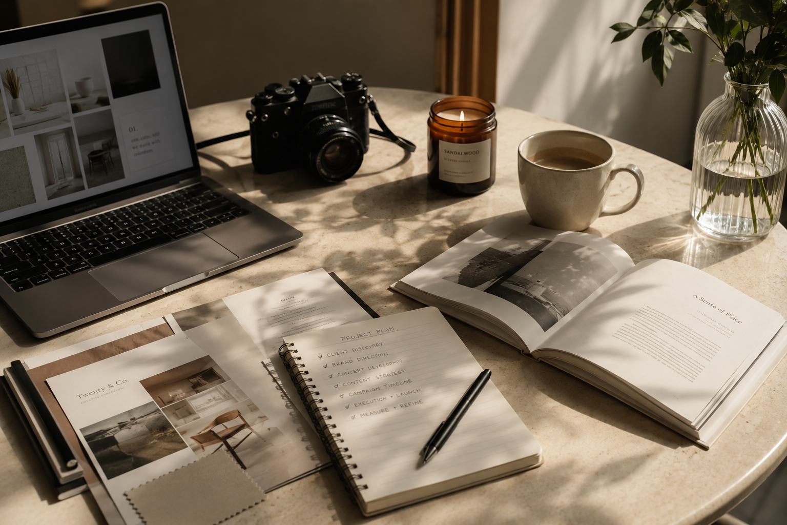 A cluttered desk with a MacBook laptop, camera, lit candle, mug, glass vase with greenery, open books, a notebook with a pen, and color swatches, all in warm natural light.