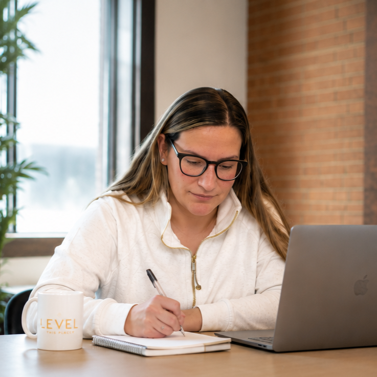 A woman sitting at a desk, writing in a notebook with a laptop in front of her and a coffee mug nearby, in an office setting.