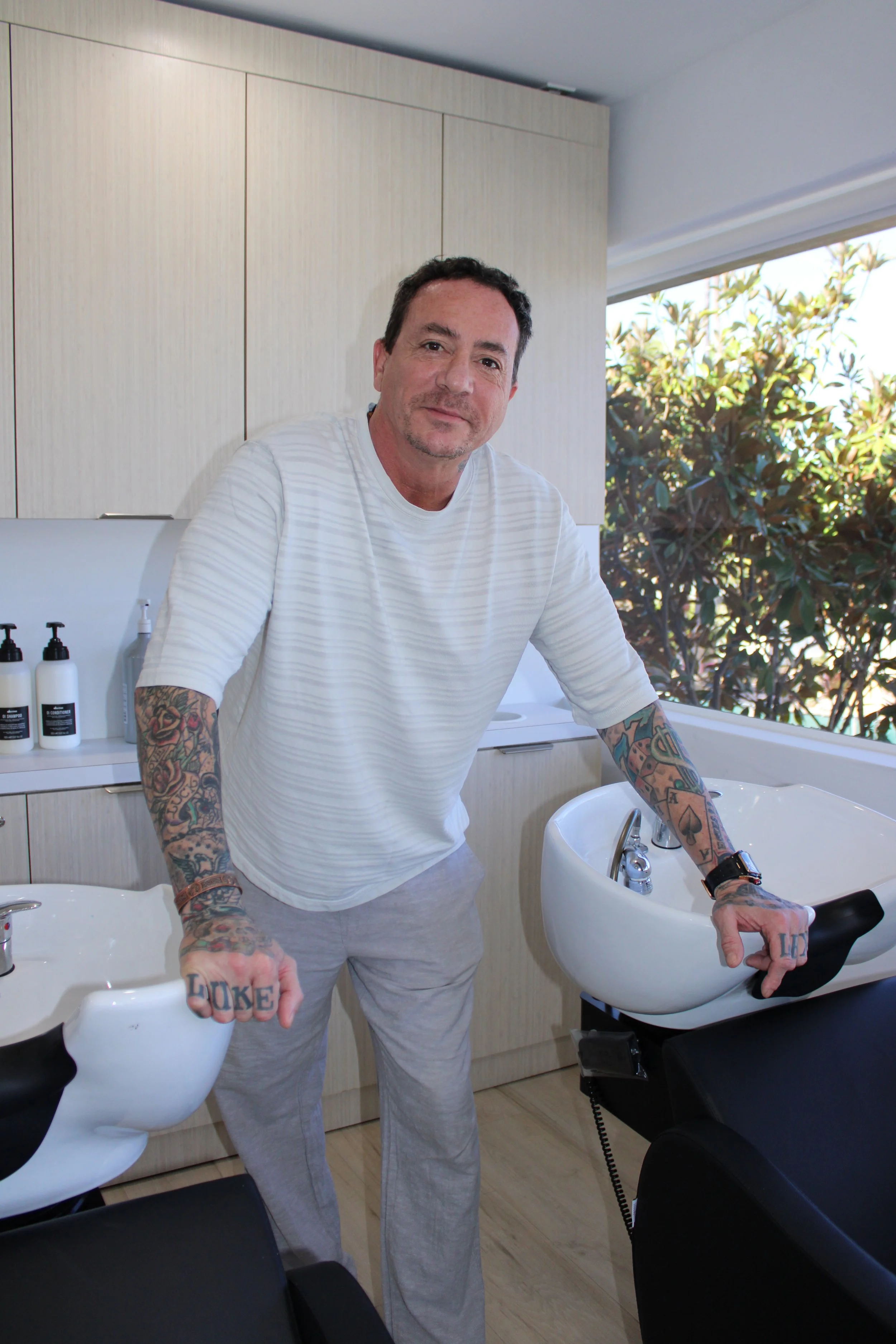 A man with tattoos on his arms and the name 'LUK' and 'DUE' tattooed on his knuckles is standing in a modern hair salon. He is wearing a white and gray striped shirt and light gray pants. The salon has light-colored cabinets, bottles of hair products on a counter, and a large window with a view of greenery outside.
