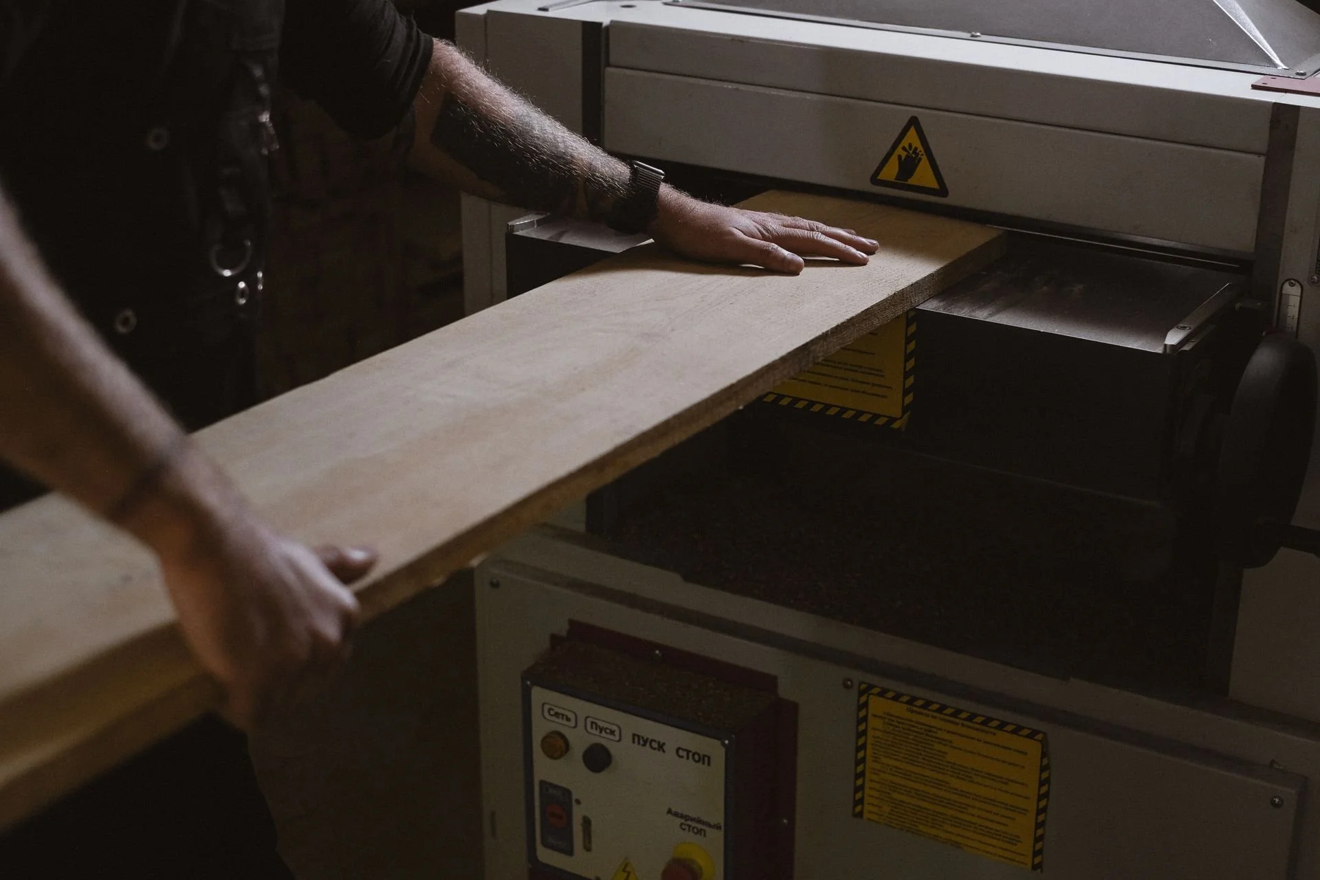 A person operating a woodworking machine, guiding a wooden board through the machine. The machine has a warning sticker and control panel with buttons and labels.