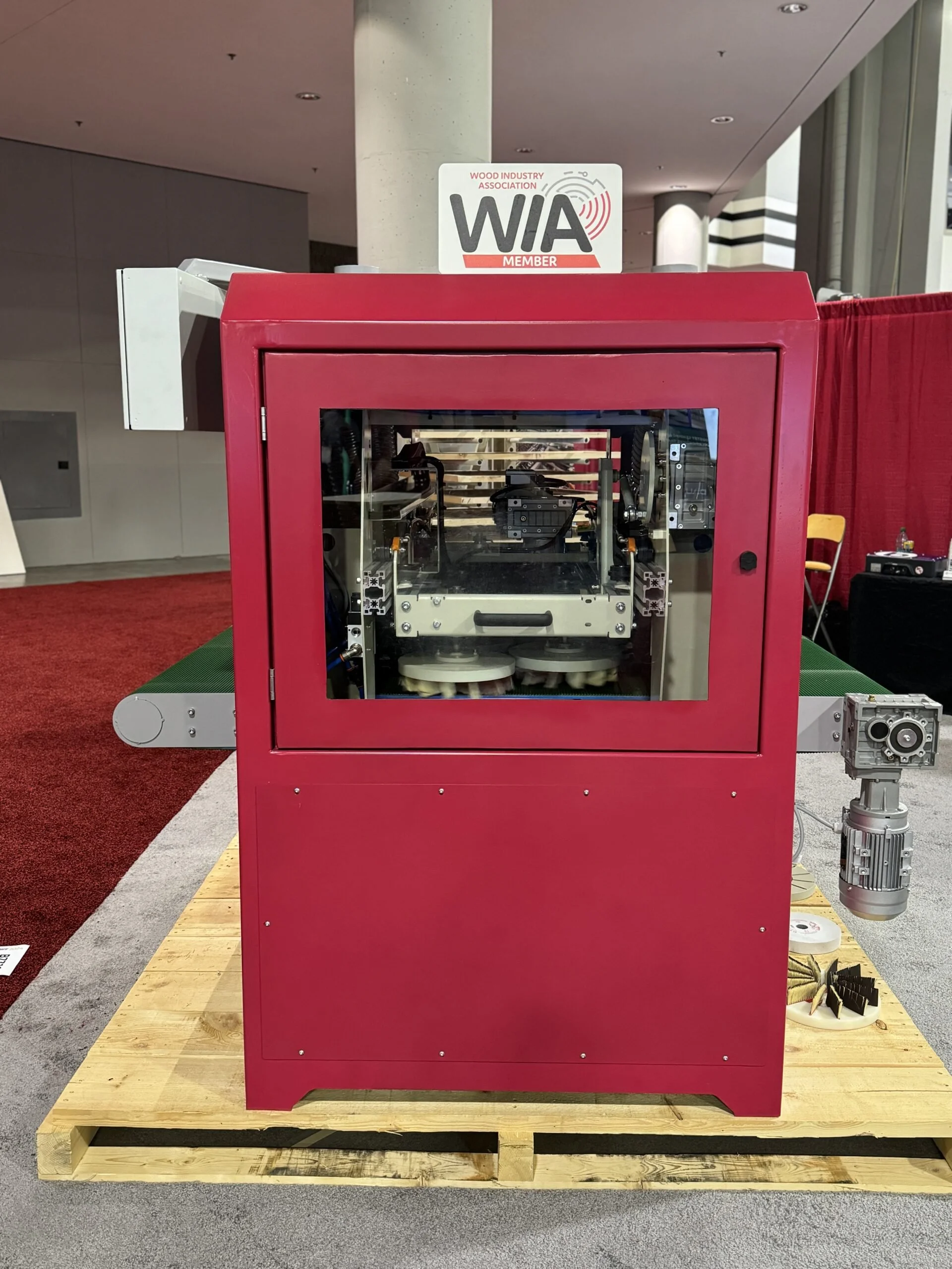 A red industrial machine with transparent glass front, placed on a wooden platform, inside a large indoor space with modern architecture. A sign on top reads 'WIA Member' of the Wood Industry Association.